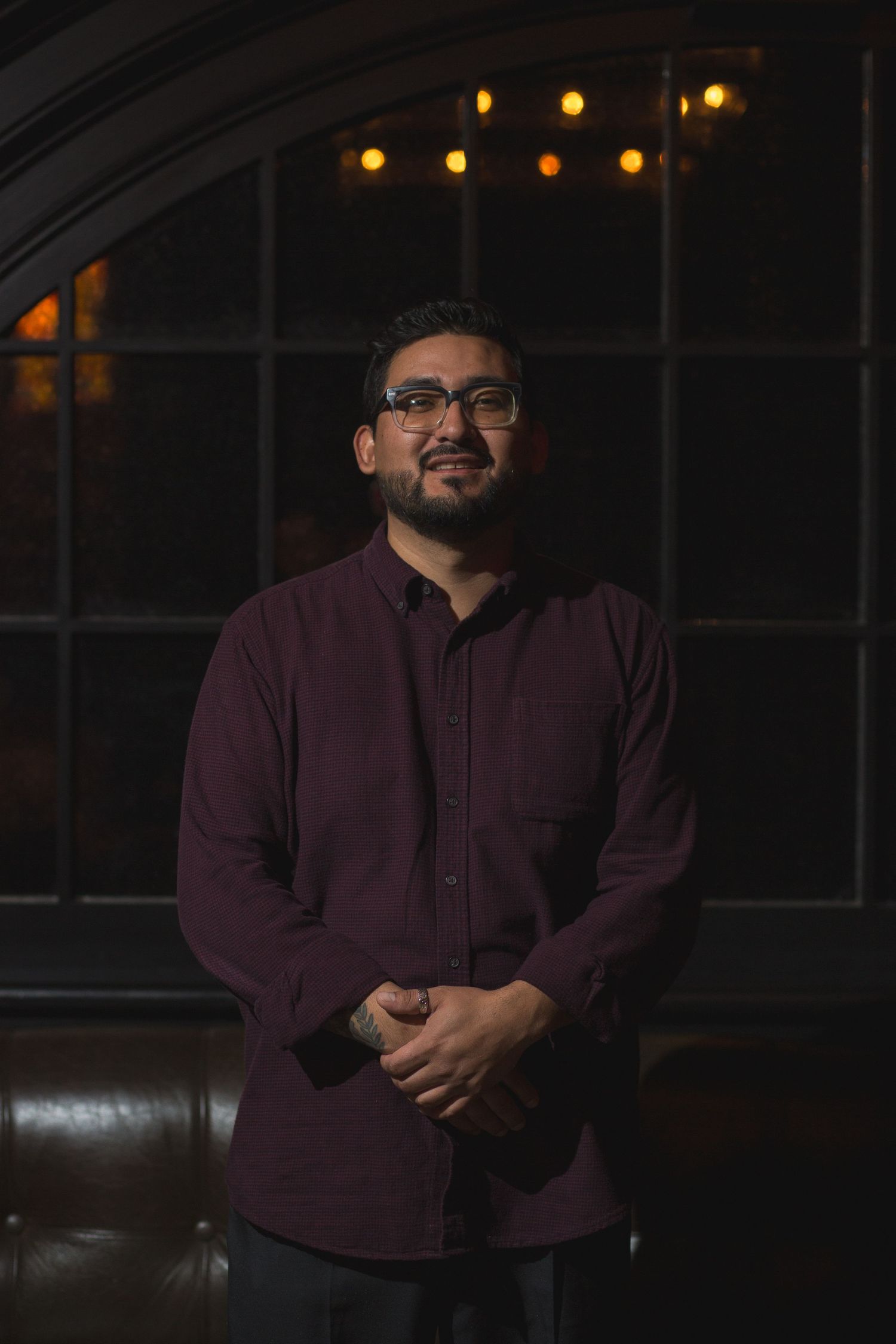 A performer in burgundy button-down shirt poses in front of an arched window with warm lighting.