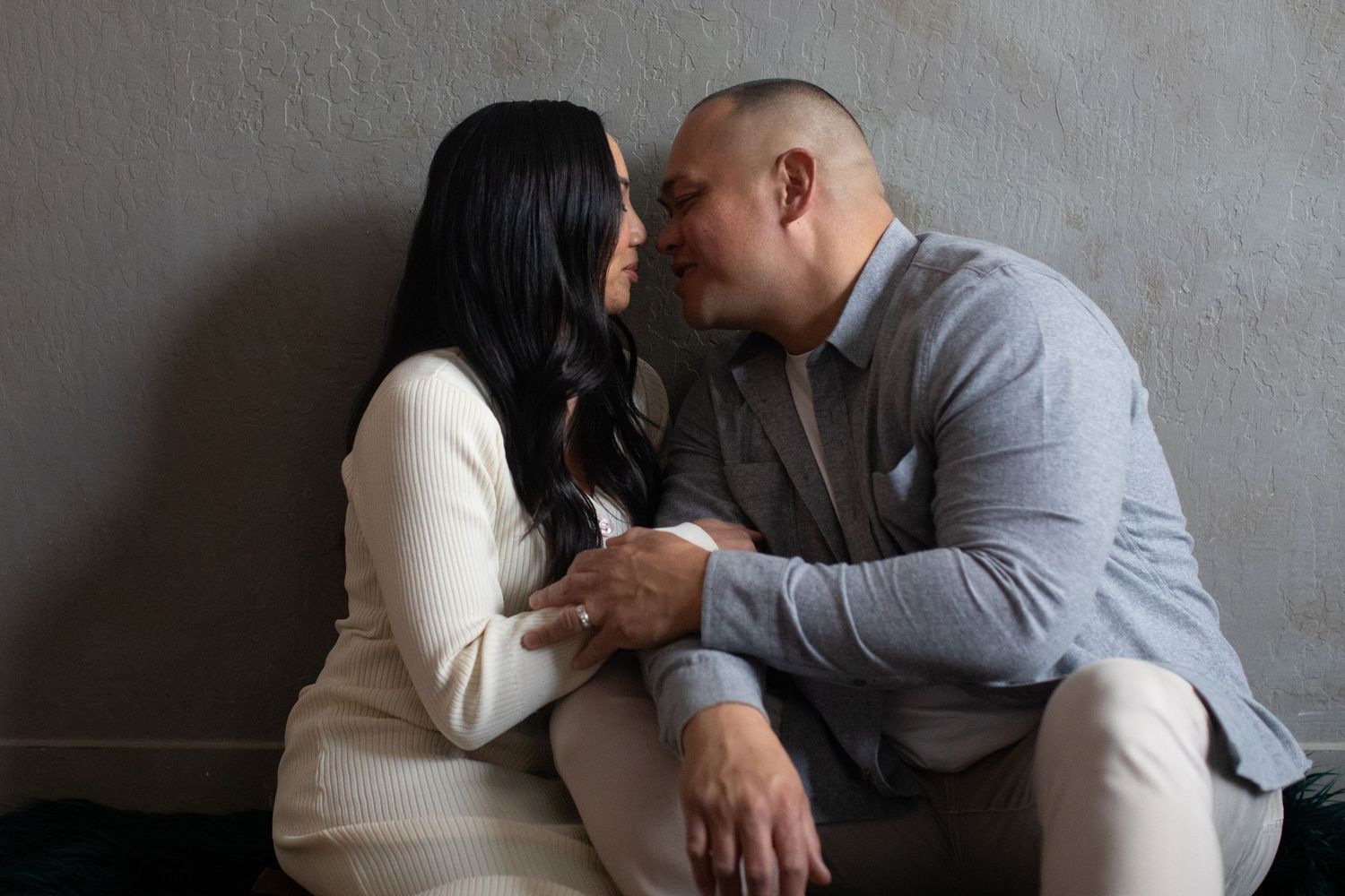 Couple in formal wear sharing intimate moment against gray wall.