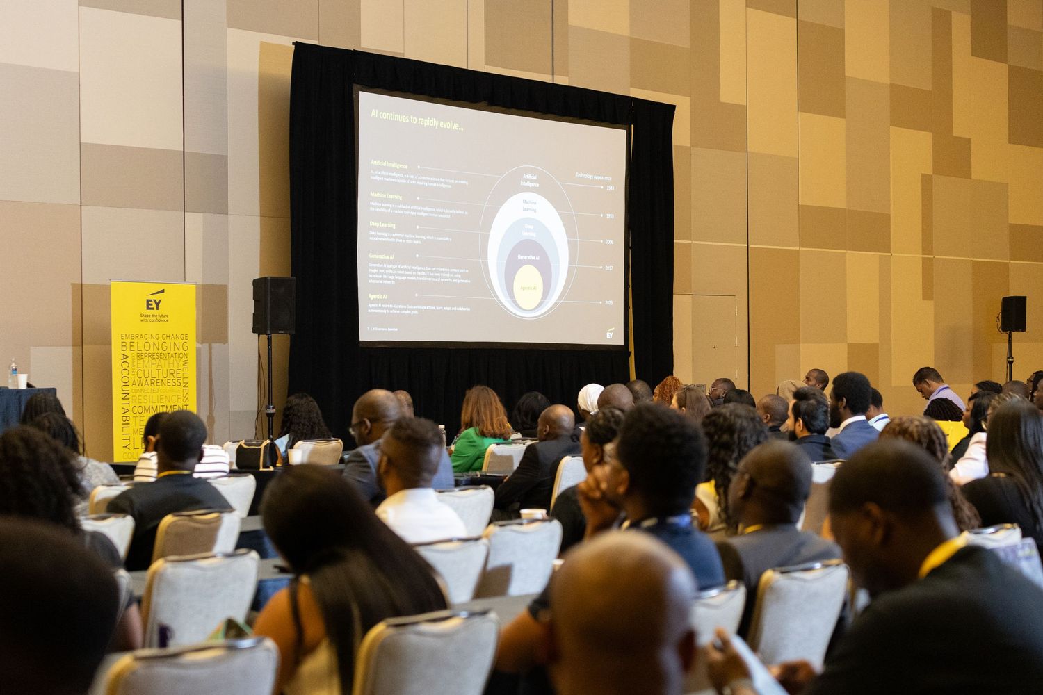 Conference attendees gather in a large ballroom with rows of chairs facing a presentation screen.
