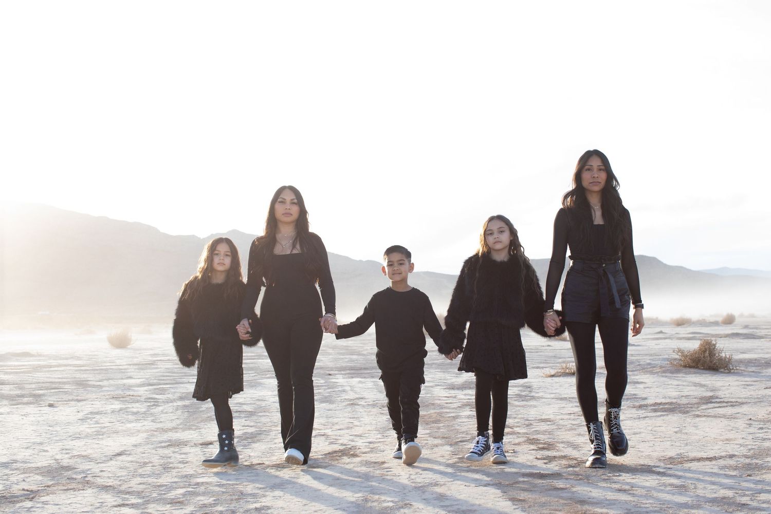 Five people in dark outfits walk hand in hand across a white desert landscape at sunset.