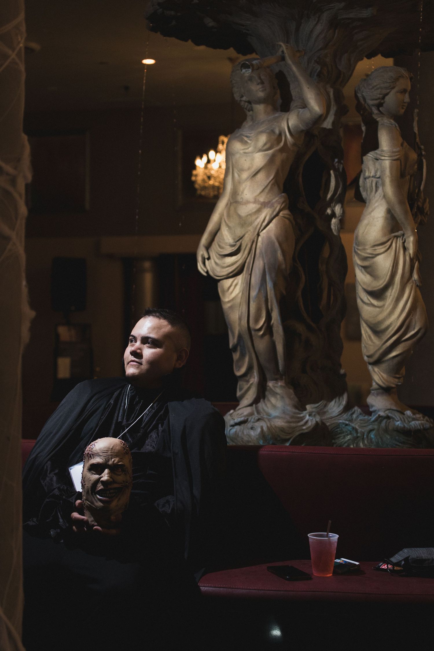 A dramatic atmospheric scene with a person in black holding a mask beside classical statues in dim lighting.