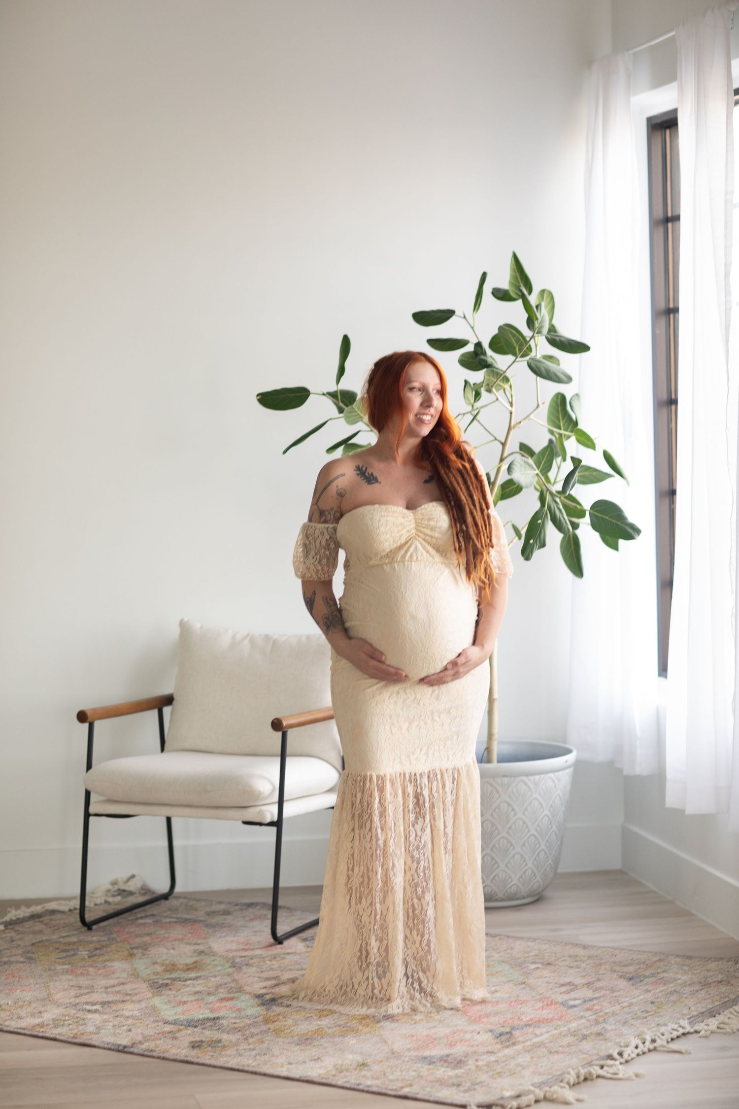 Pregnant woman in cream dress poses elegantly by indoor plant and sheer curtains.