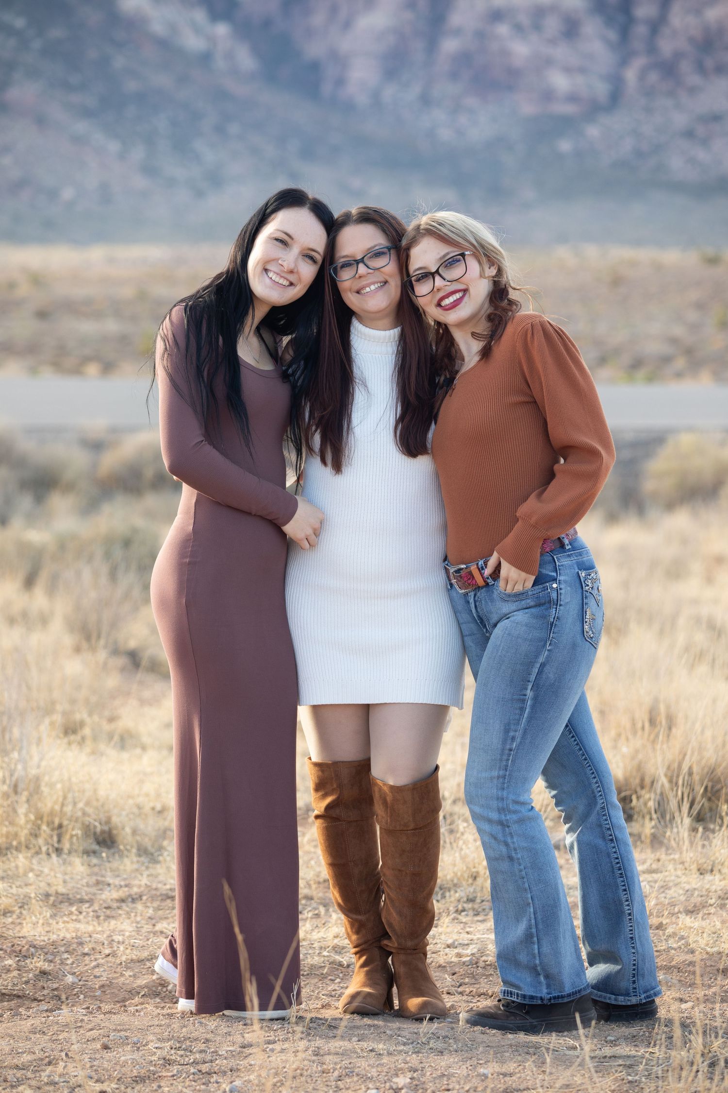 Three friends pose together in stylish outfits against a desert mountain landscape.