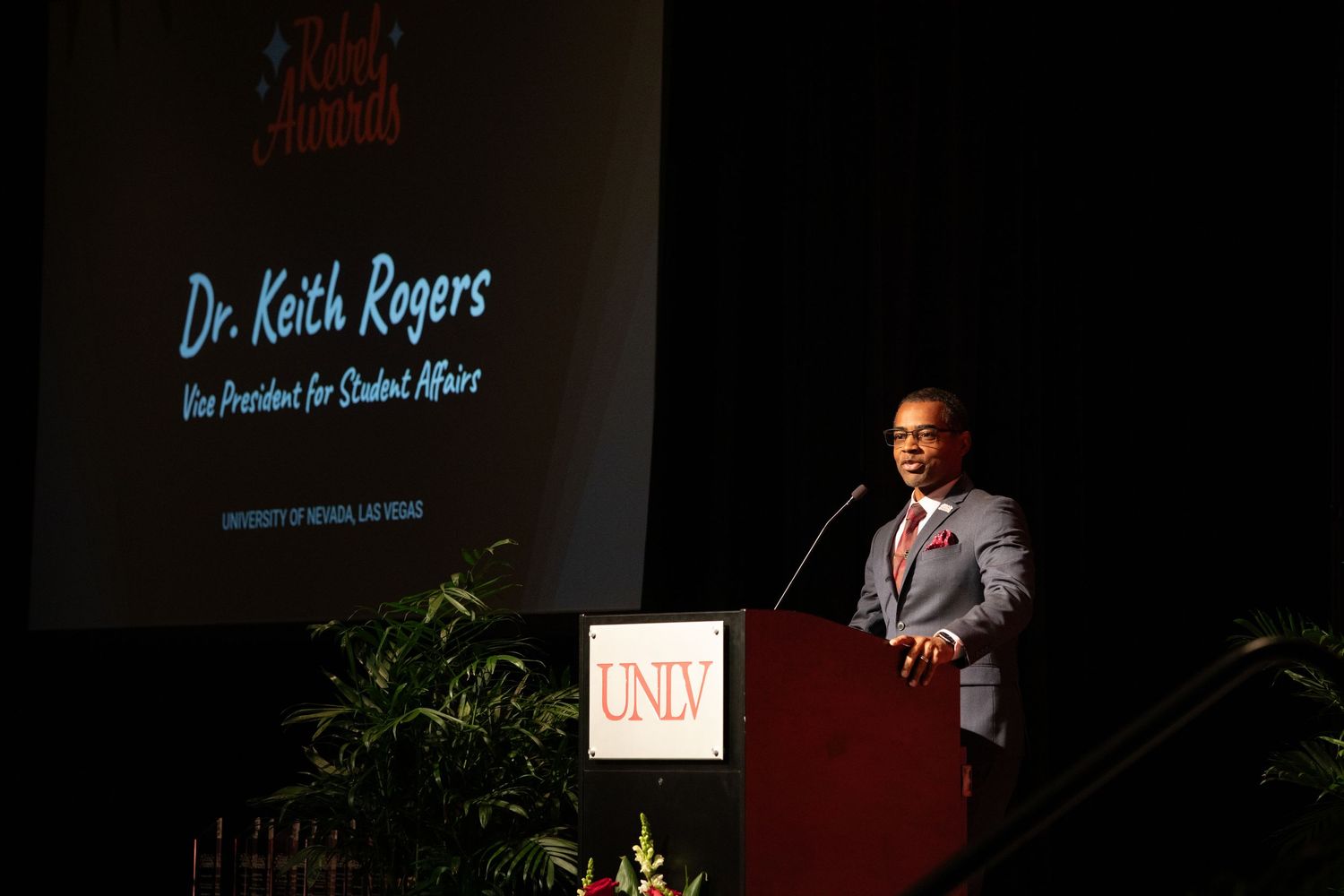 Speaker presents at UNLV podium with projection screen displaying text in background.