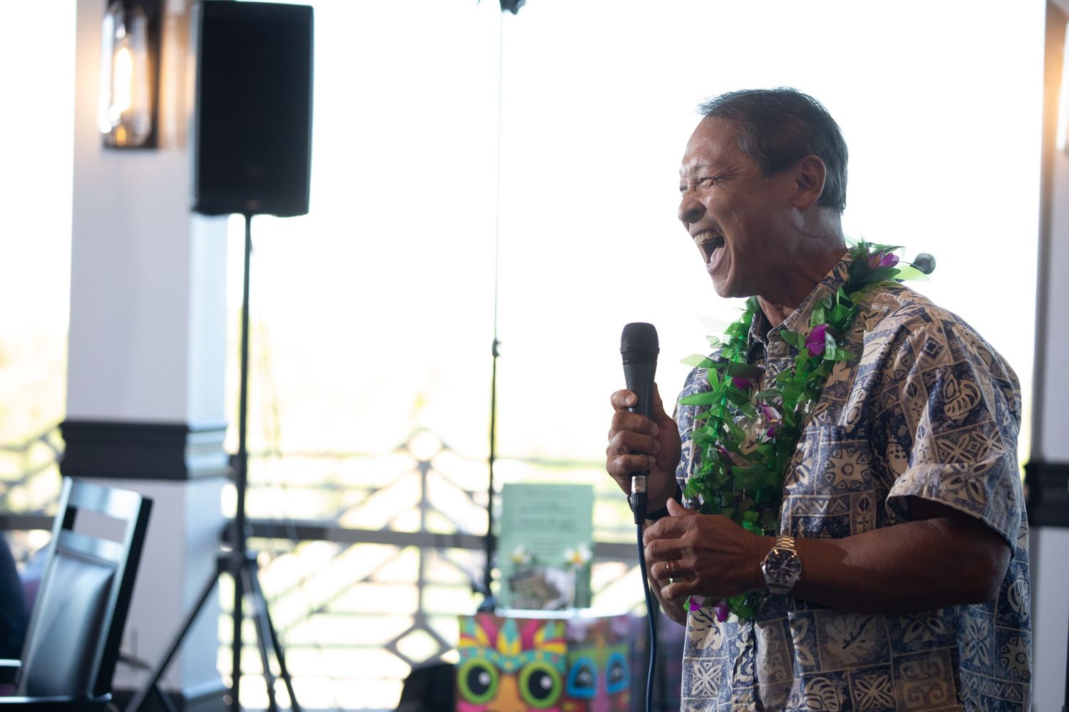Person speaking into microphone wearing Hawaiian shirt at party with scenic background.