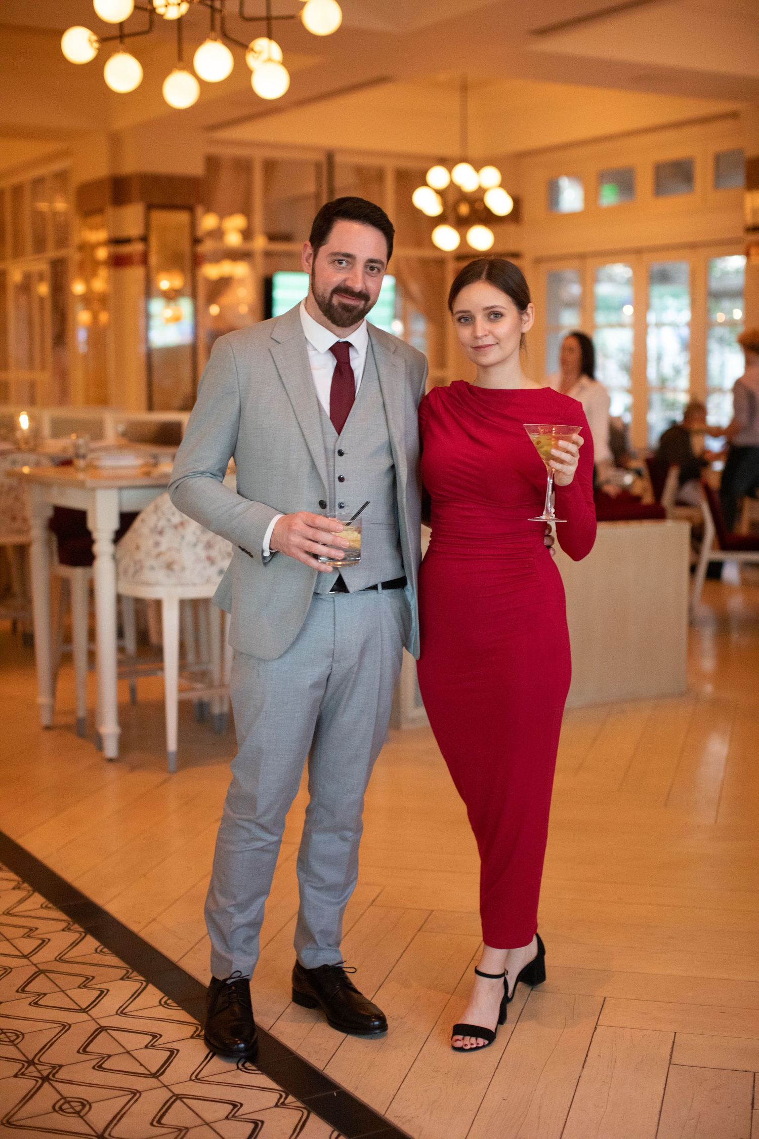 A couple dressed in grey suit and red dress pose together at an elegant venue with warm lighting.