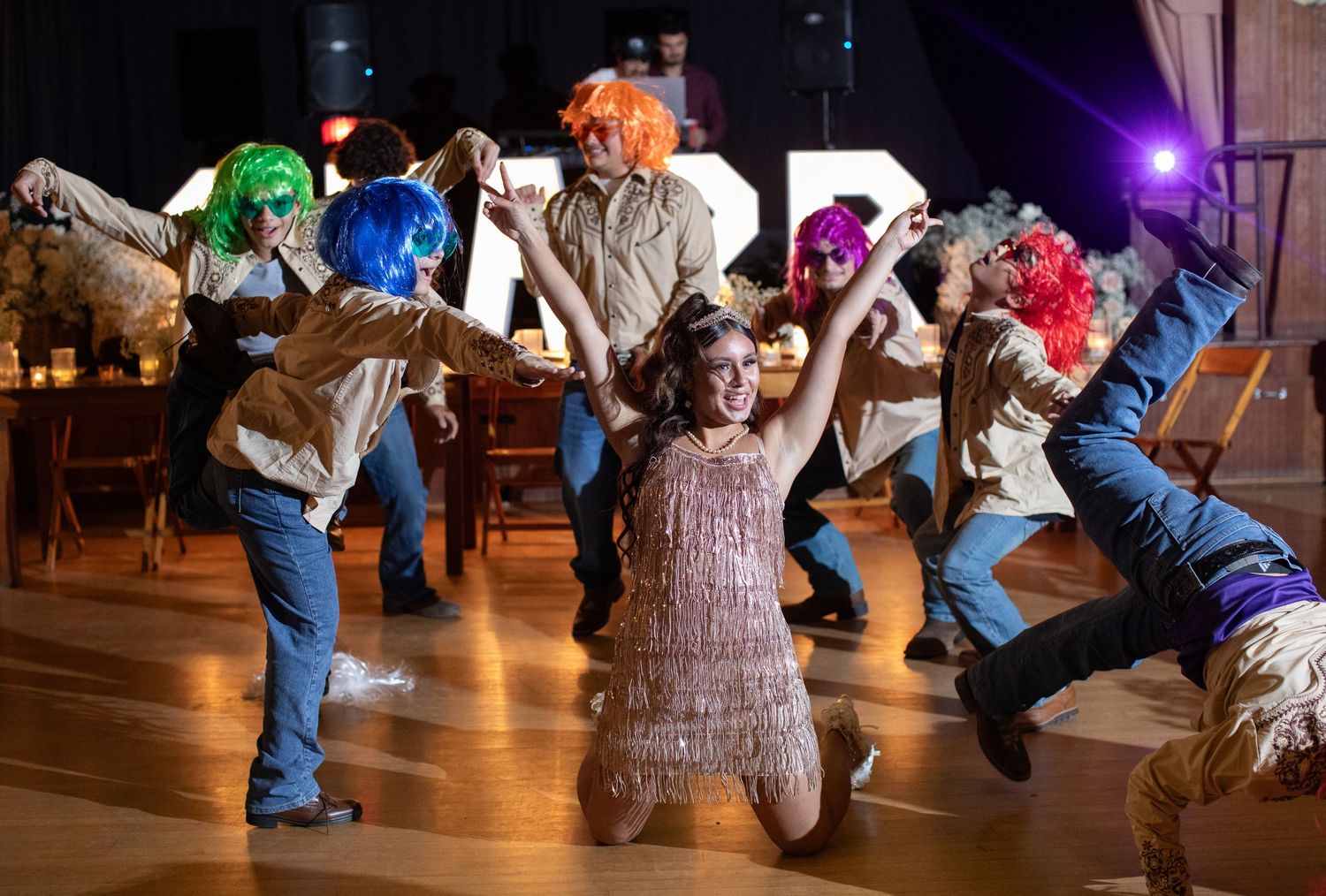 Costumed dancers performing energetic choreography with colorful wigs on a wooden dance floor.