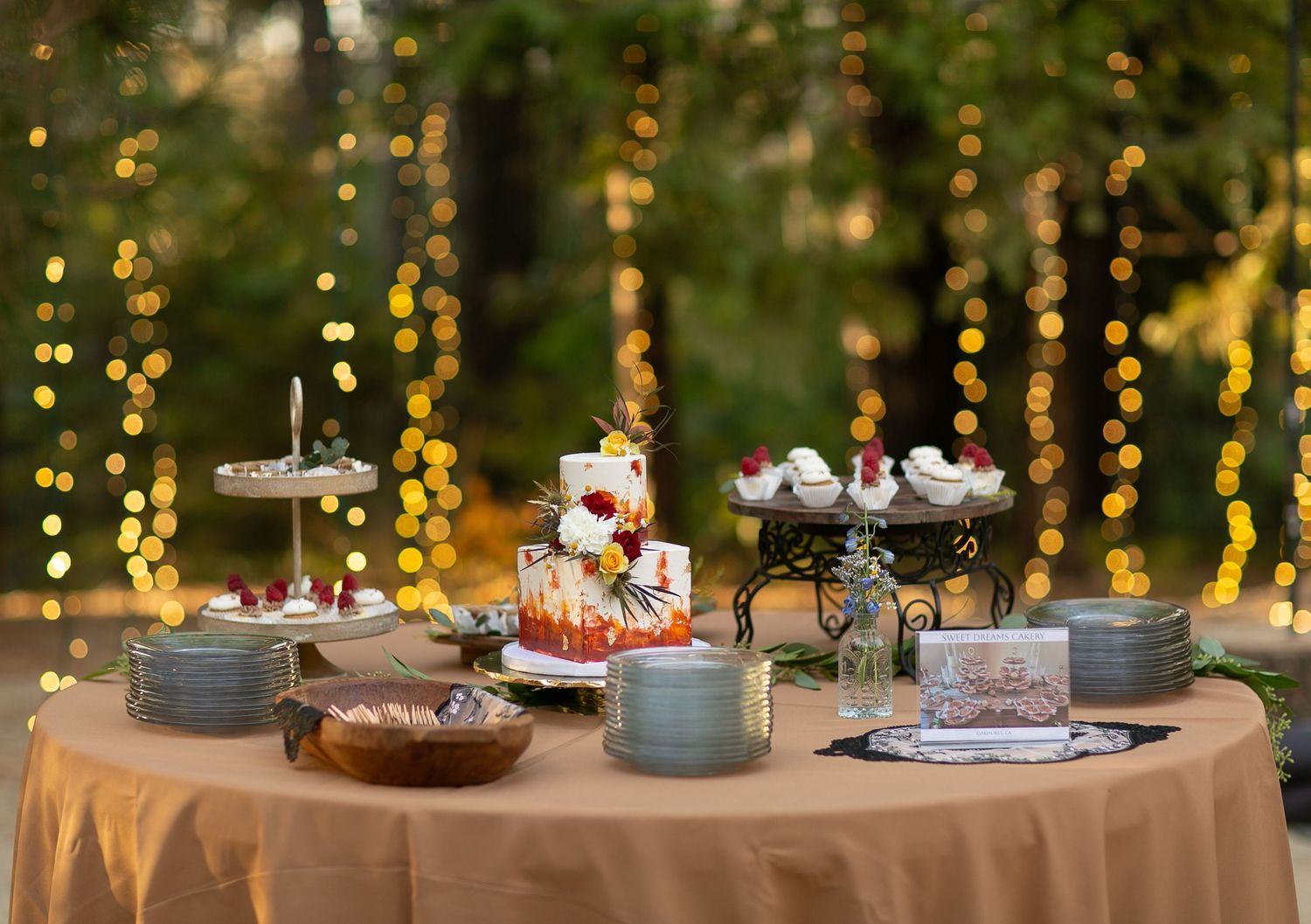 Dessert table with cupcakes and treats decorated with string lights in outdoor setting.