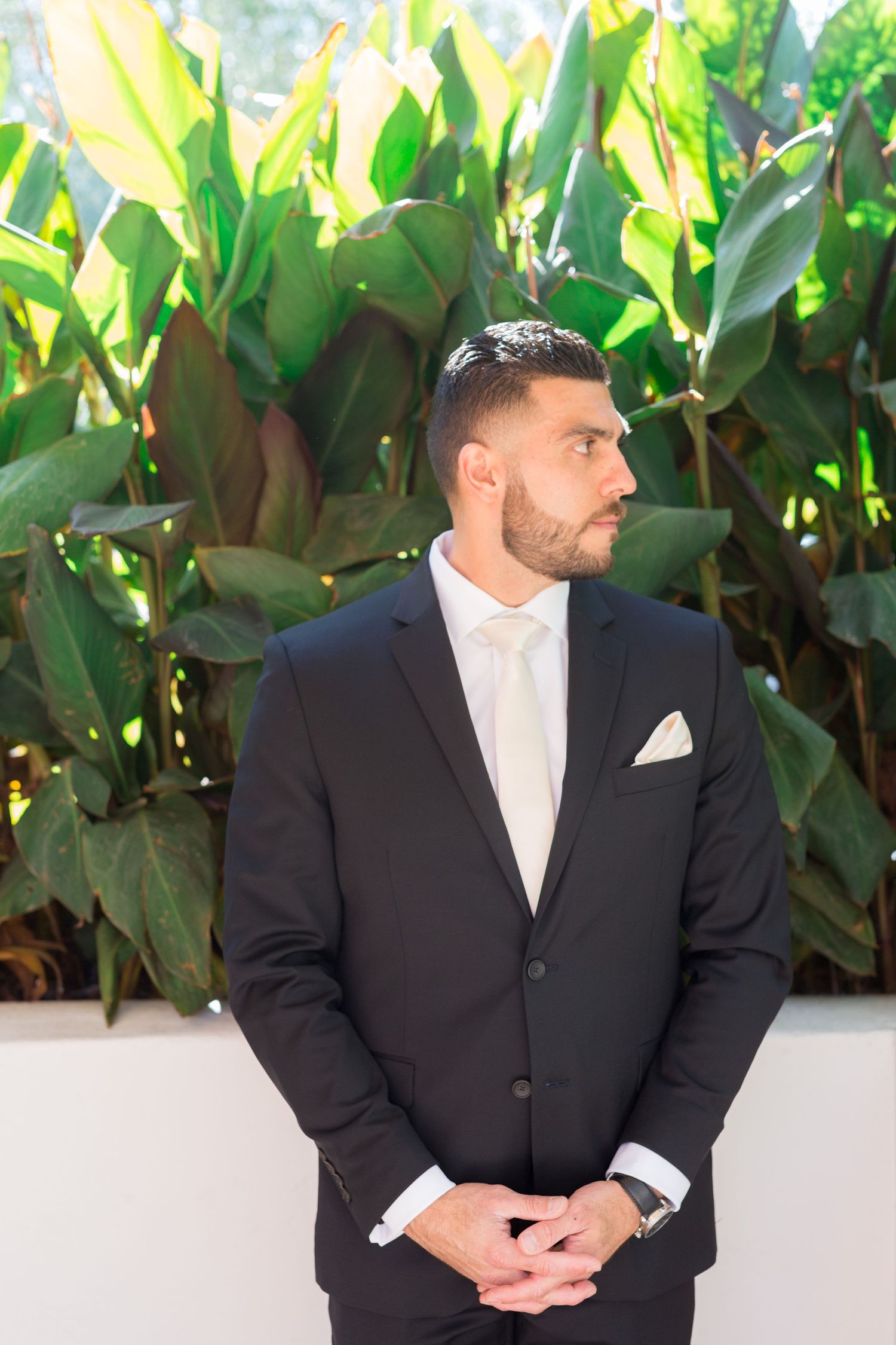 A groom in a classic black suit stands against a backdrop of tropical foliage.
