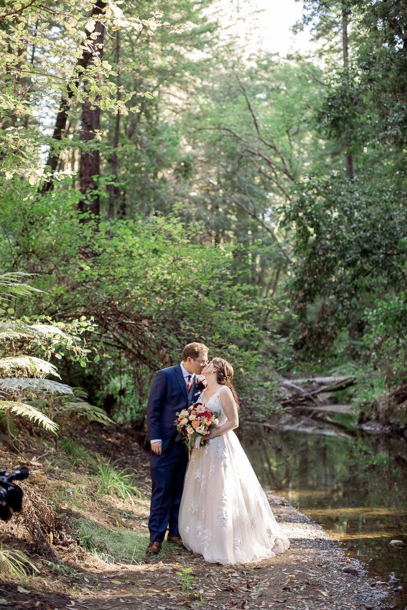 A romantic wedding photo captured in a lush forest setting beside a tranquil stream.