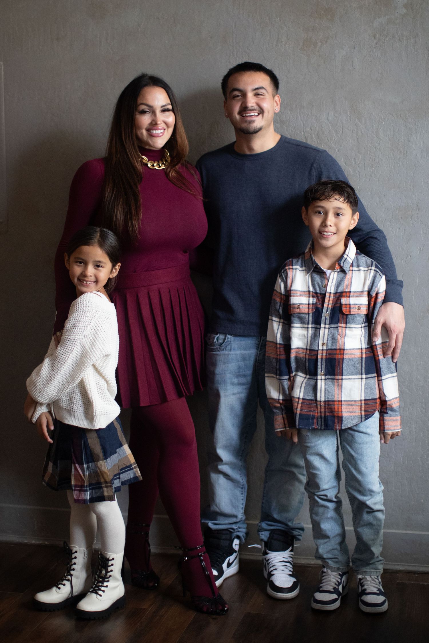 Family portrait series against gray wall with coordinating burgundy dress and plaid shirt outfits.