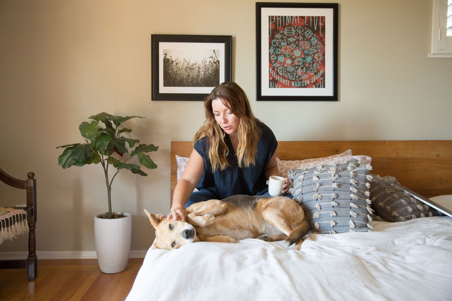 Woman relaxes on bed with golden retriever while holding coffee cup near wall artwork and potted plant.