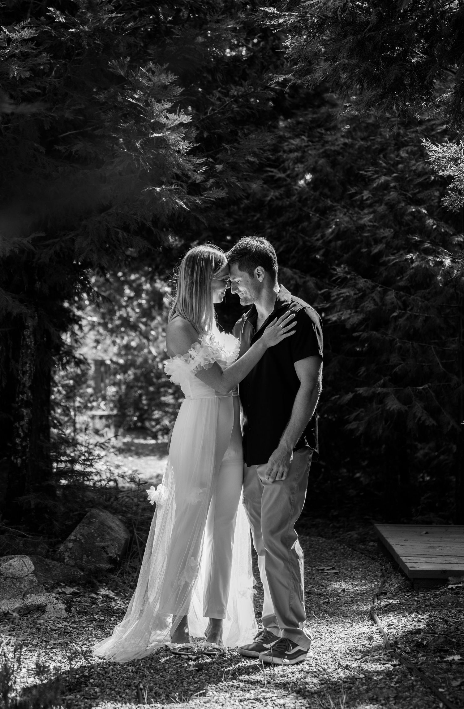 A black and white photo of a couple sharing an intimate moment in a wooded setting.