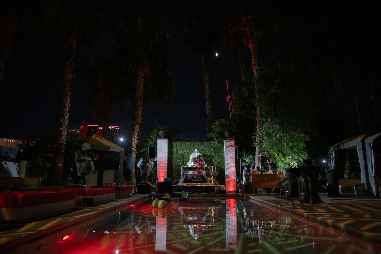 Nighttime outdoor scene with palm trees and illuminated pillars reflected in wet pavement.