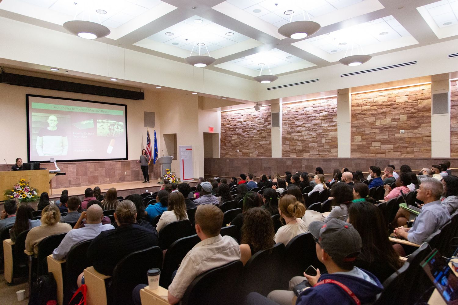 A full auditorium of attendees watching a presentation on a large screen in a lecture hall.