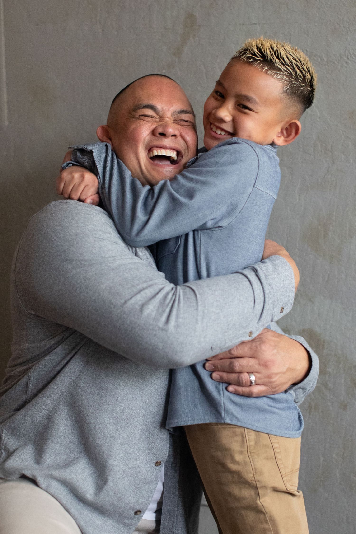 Father and son sharing joyful embrace and laughter against gray wall.