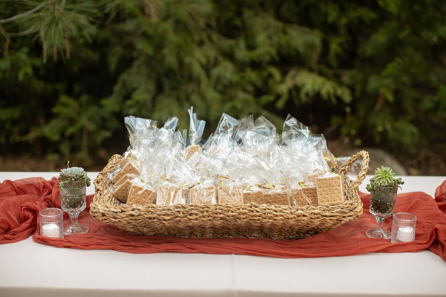Glass favor bottles arranged in a woven basket on a red tablecloth at an outdoor event.
