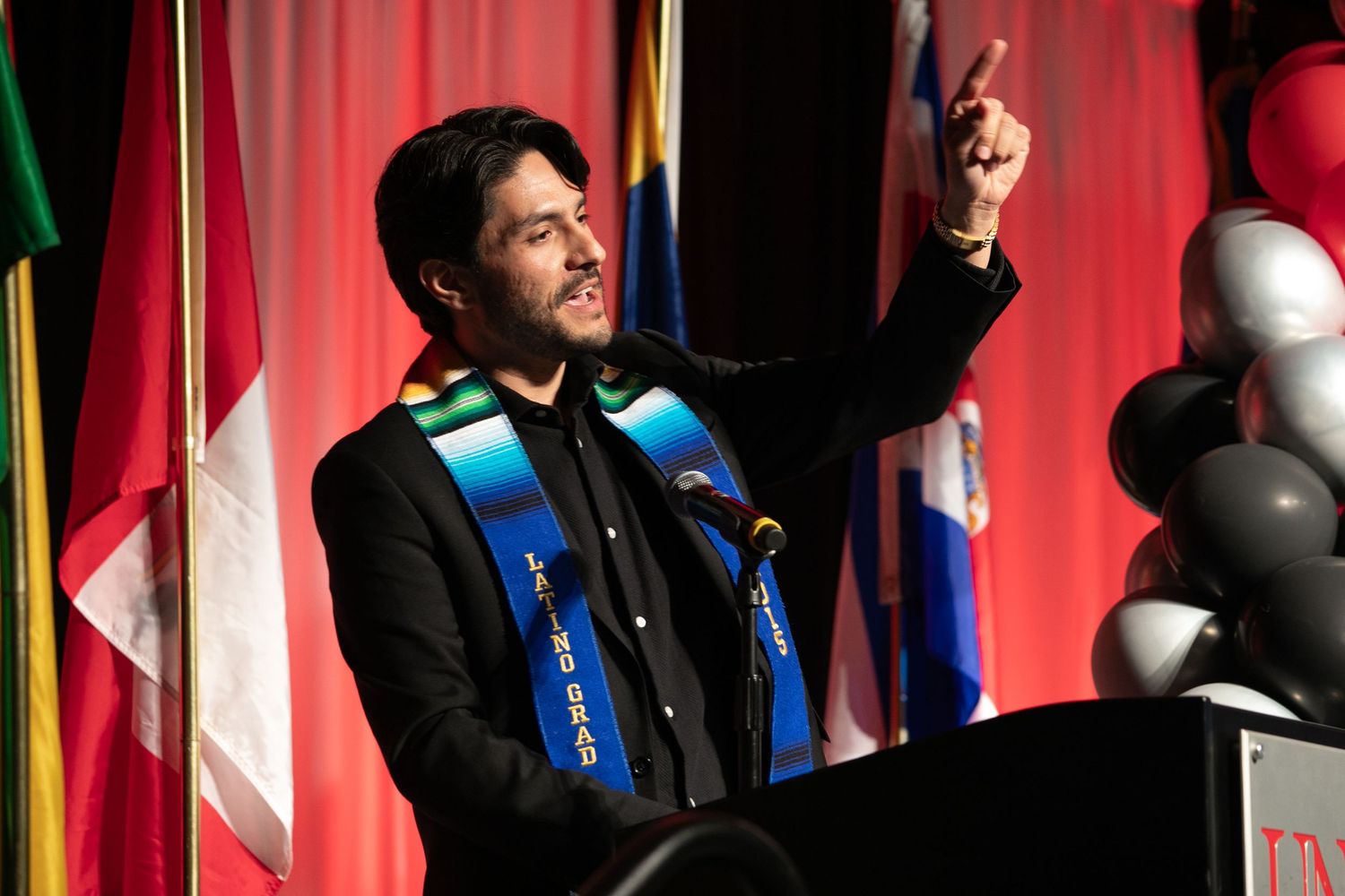 Speaker holds microphone at podium with red curtain and balloons in background.