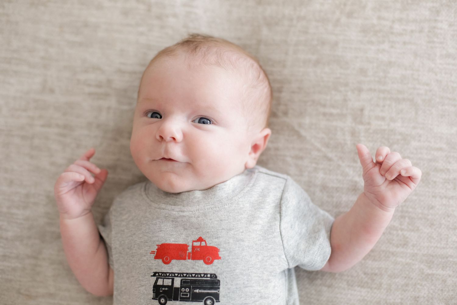Baby lying on light gray fabric wearing a simple gray t-shirt with arms outstretched.