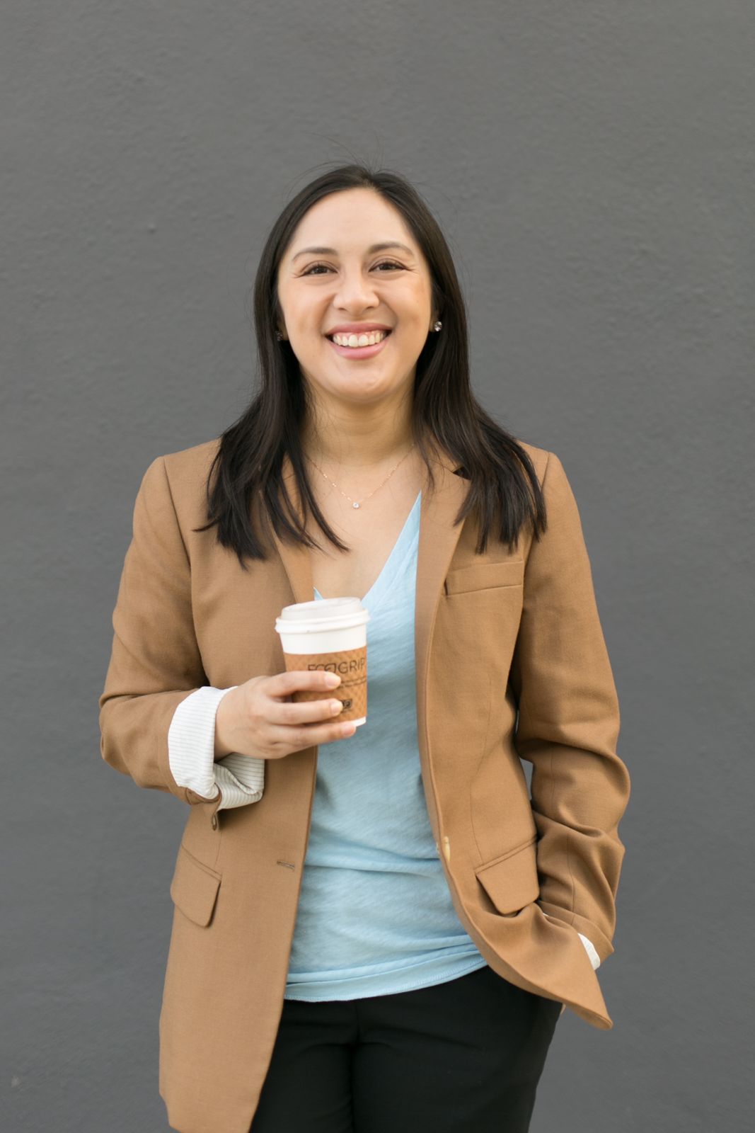 Professional in tan blazer holding coffee cup against grey wall background.