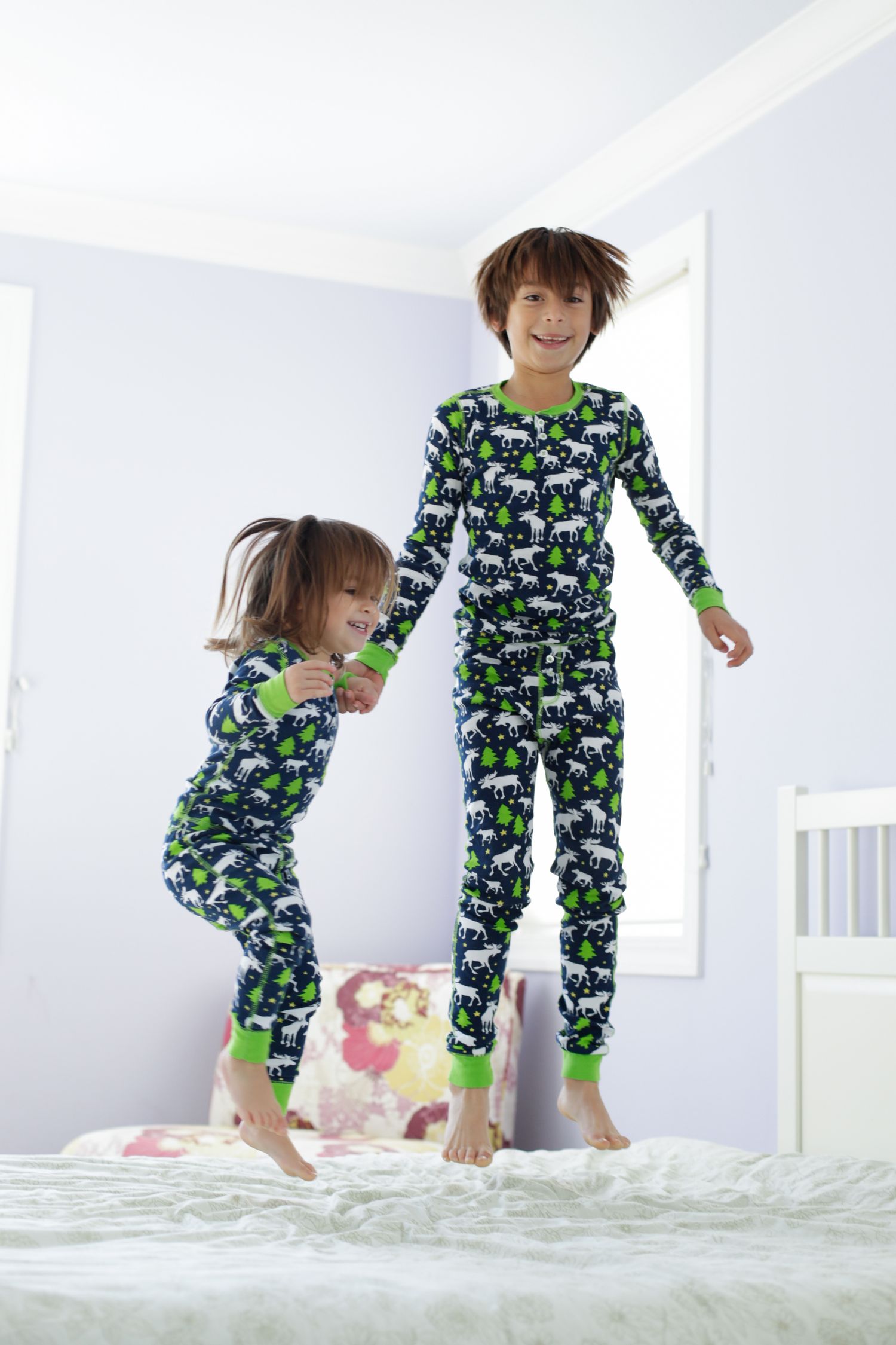 Two children in matching dinosaur print pajamas jumping on a white bed in a bright room.