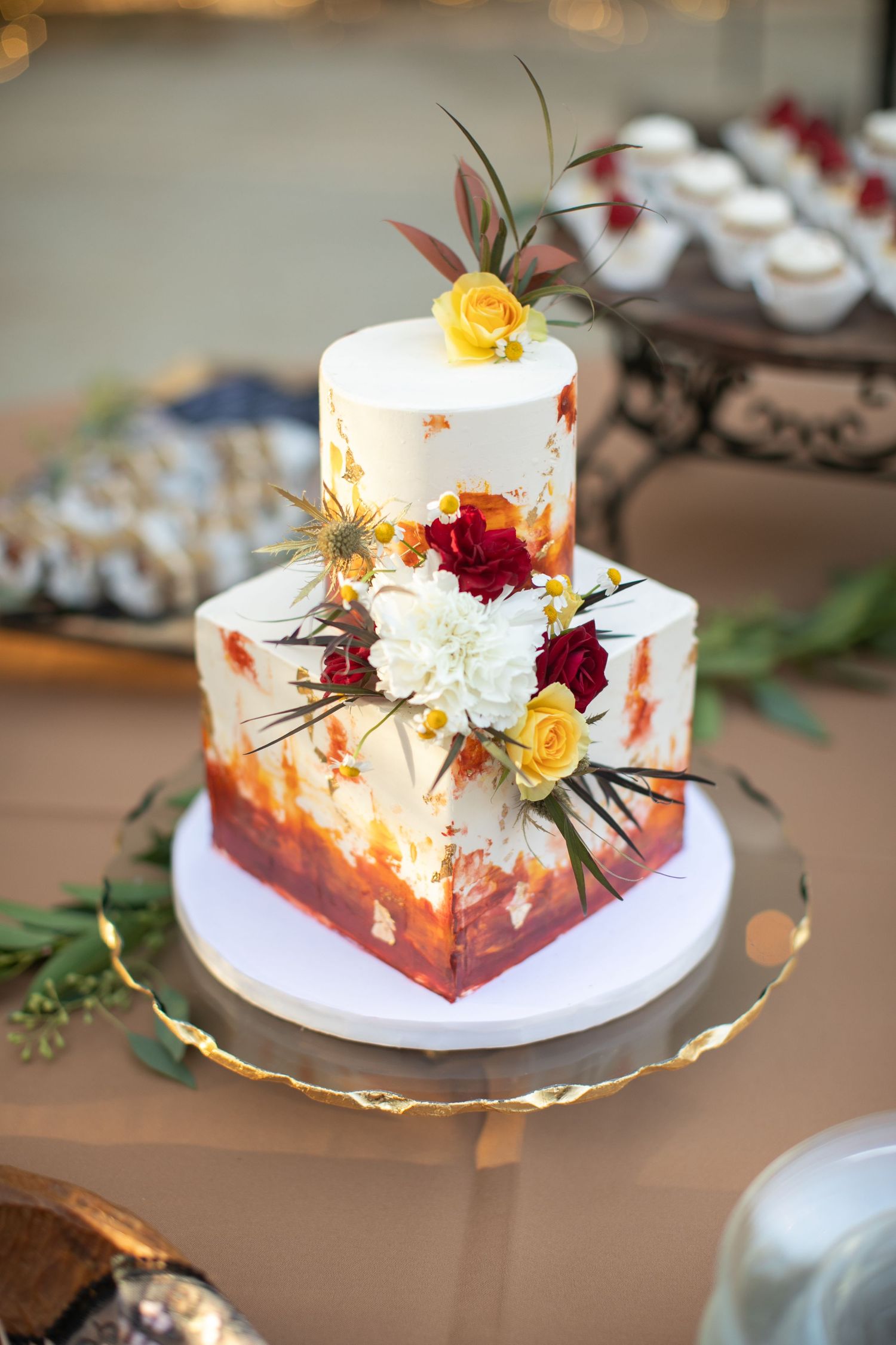 A white wedding cake decorated with burgundy and yellow flowers on a gold cake stand.