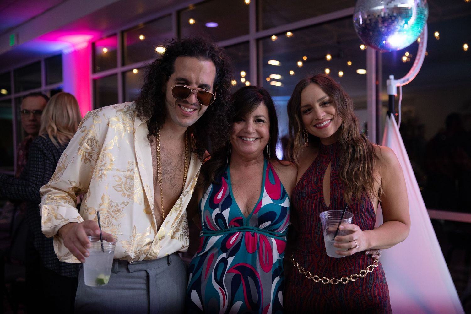 Party guests socialize and pose together under magenta lighting at a nighttime celebration.