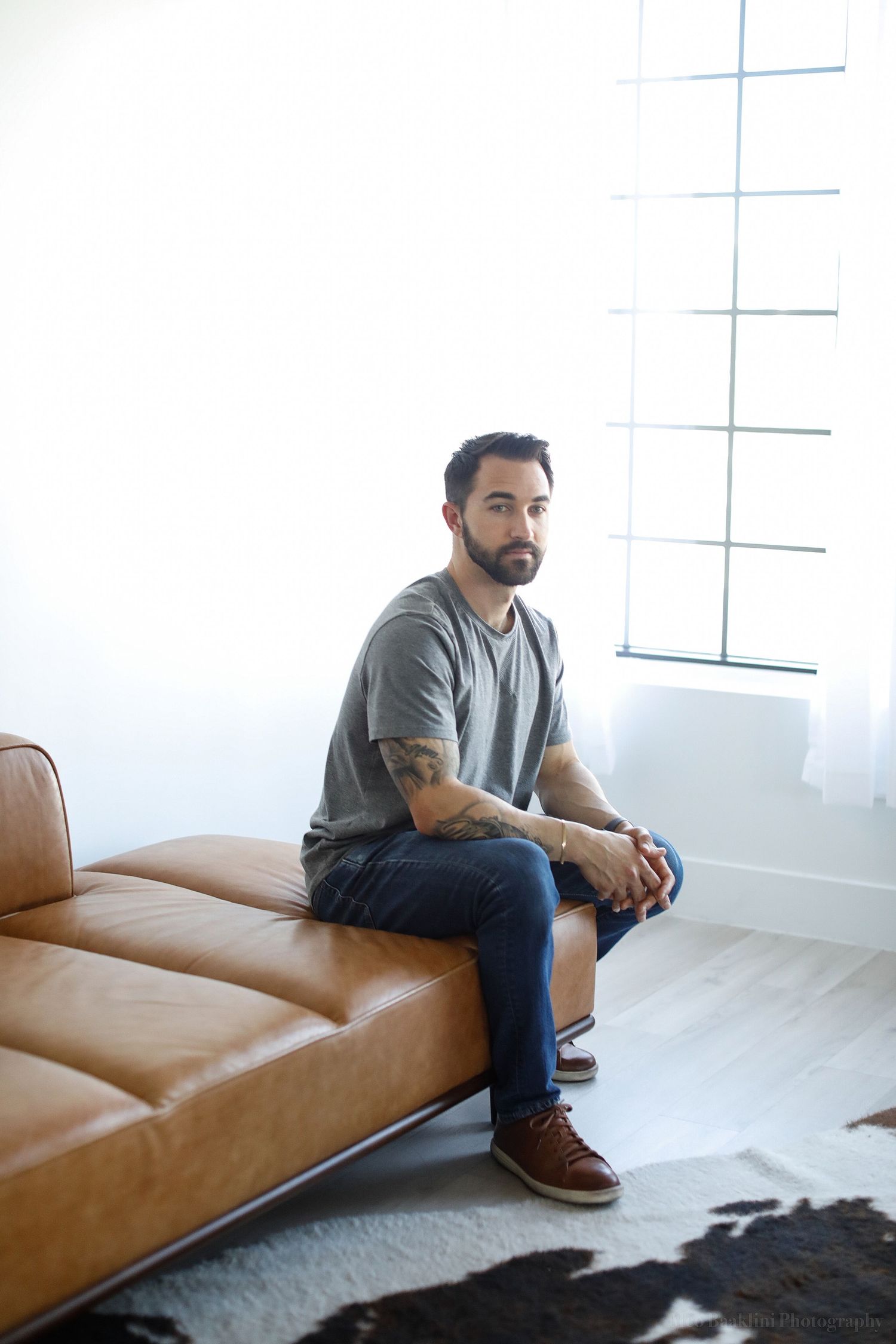 Person in grey shirt sits contemplatively on brown leather couch near bright window.
