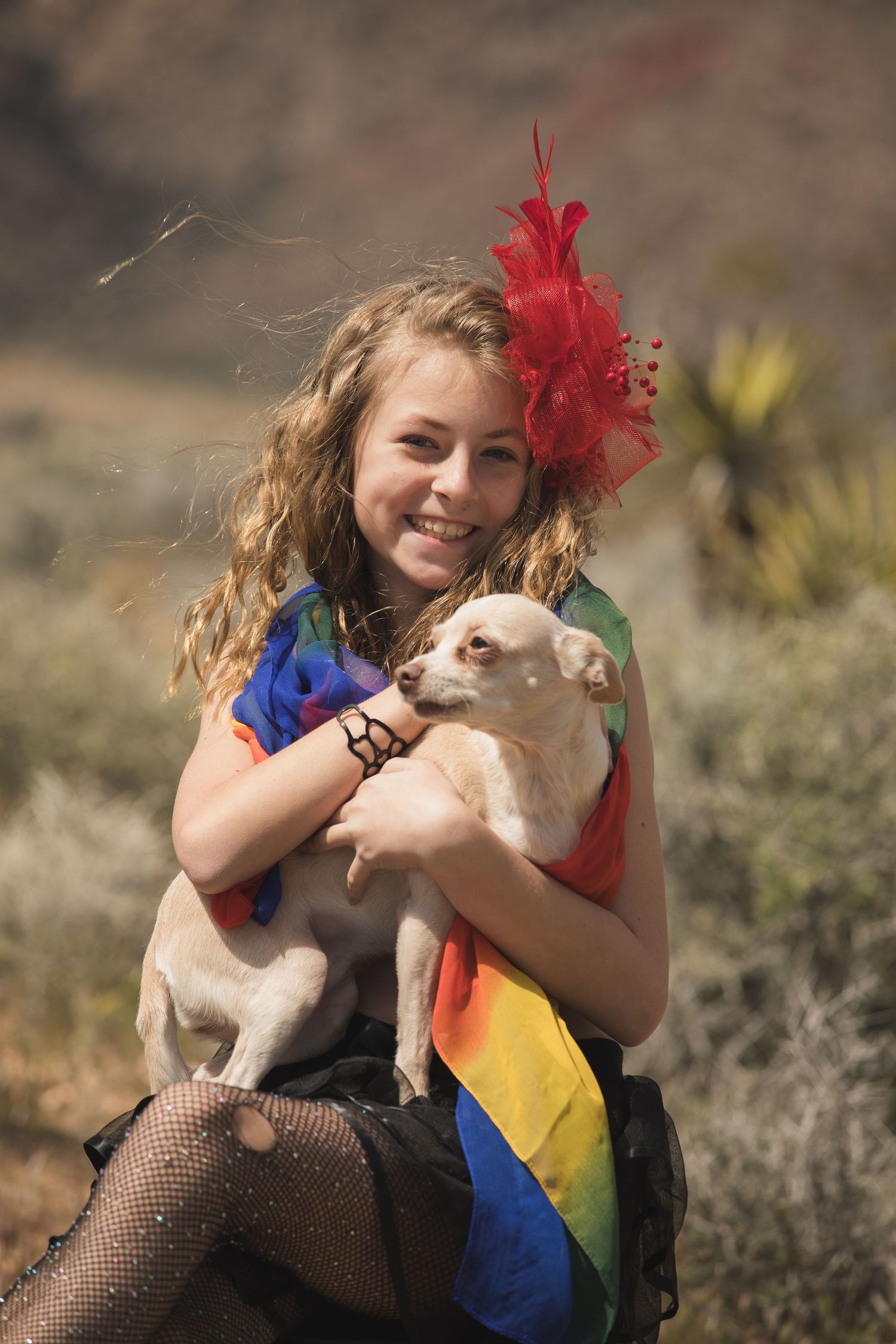 Rainbow-dressed figure holds small dog while posing in desert landscape with red feather accessory.