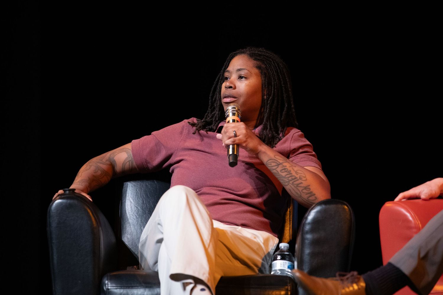 Speaker in a burgundy shirt sits casually during panel discussion on dark stage.