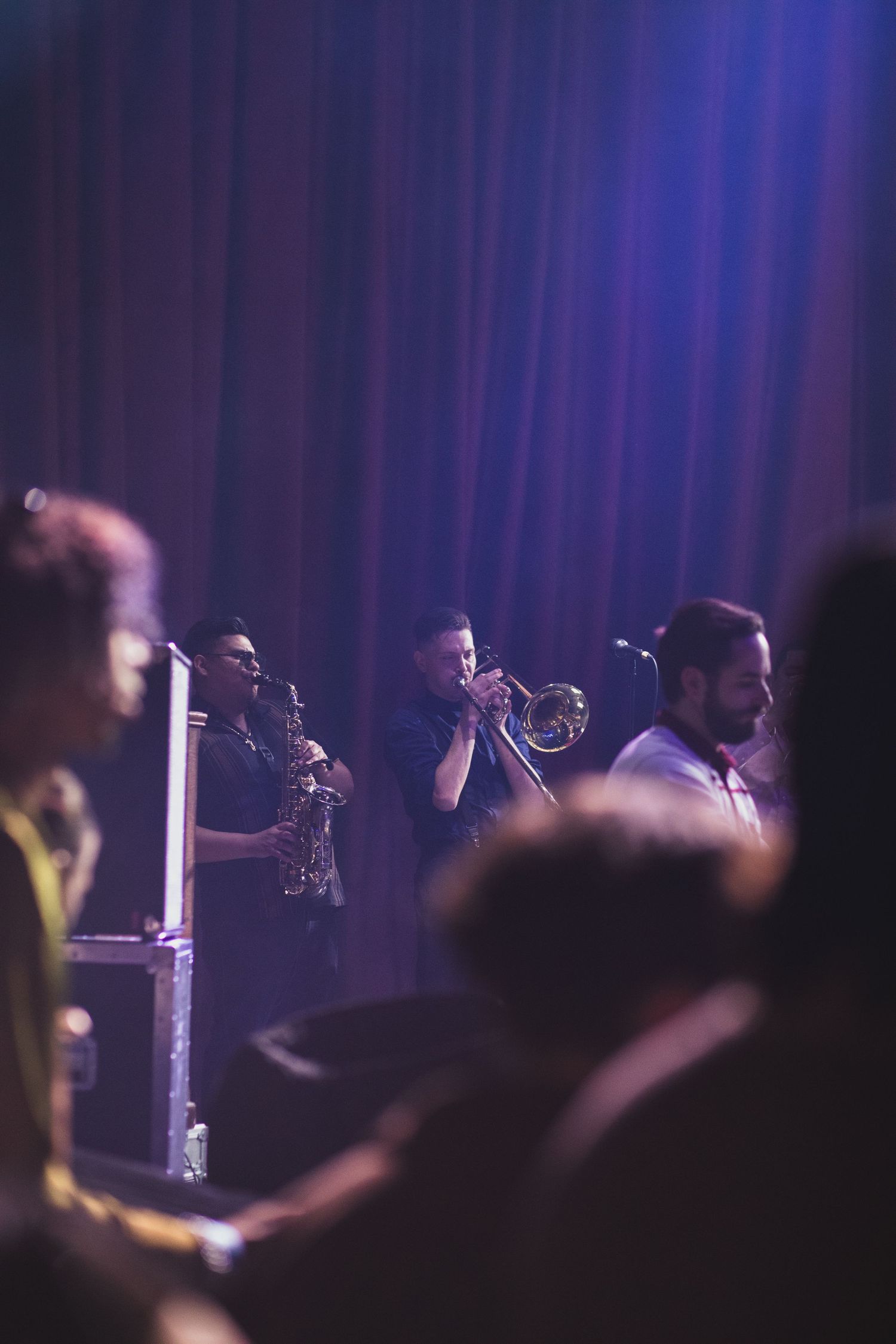 Horn section musicians perform on stage with dramatic purple stage lighting and curtain backdrop.