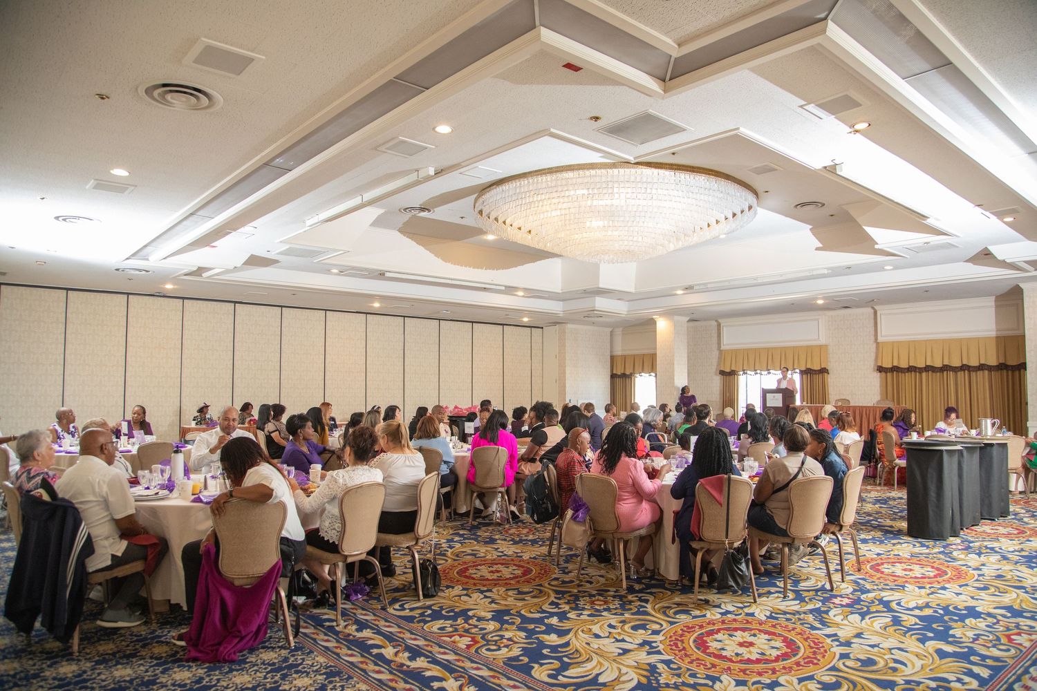 Large conference room with ornate ceiling and carpet filled with attendees seated at round tables.