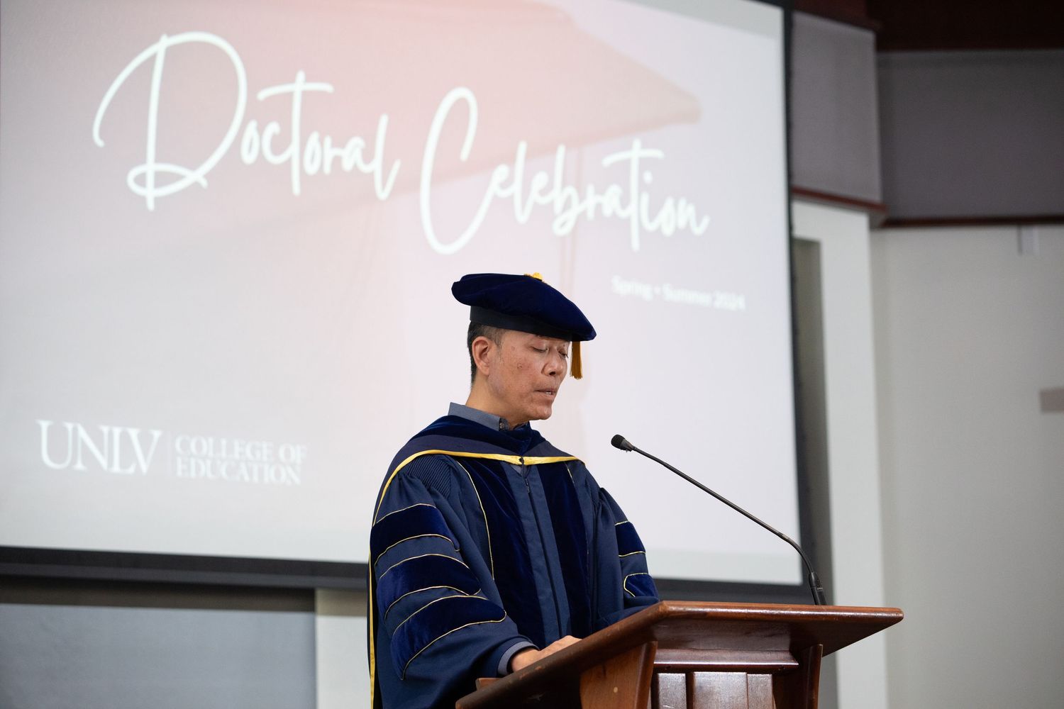 Academic speaker in doctoral regalia delivers remarks at a podium during UNLV graduation ceremony.