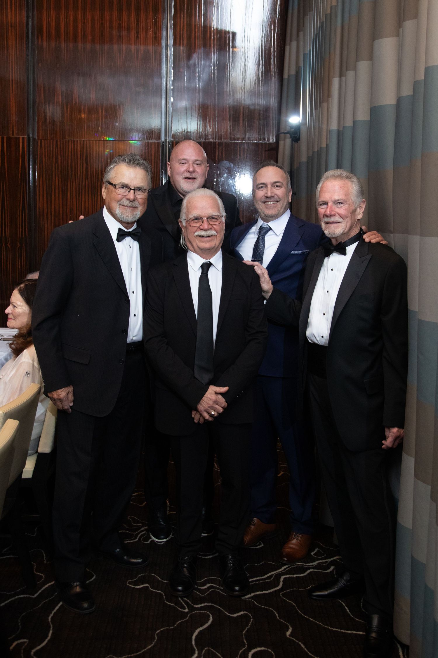A group in formal attire and tuxedos pose together against a dark background.