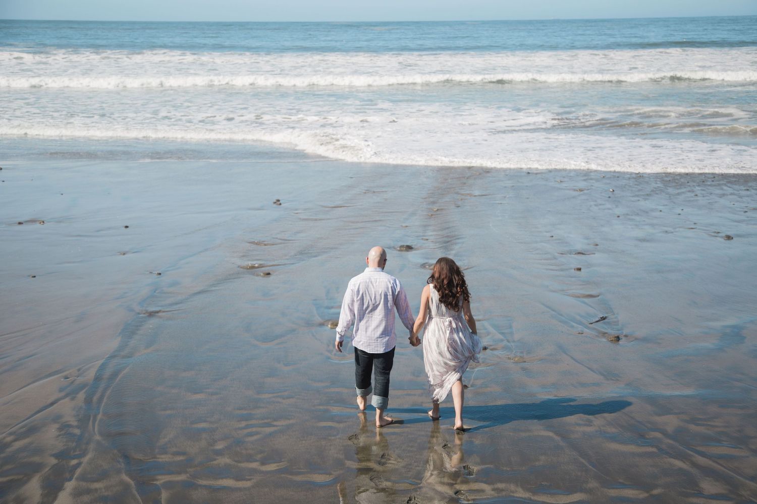 Couple walking hand in hand along wet sand at ocean's edge.
