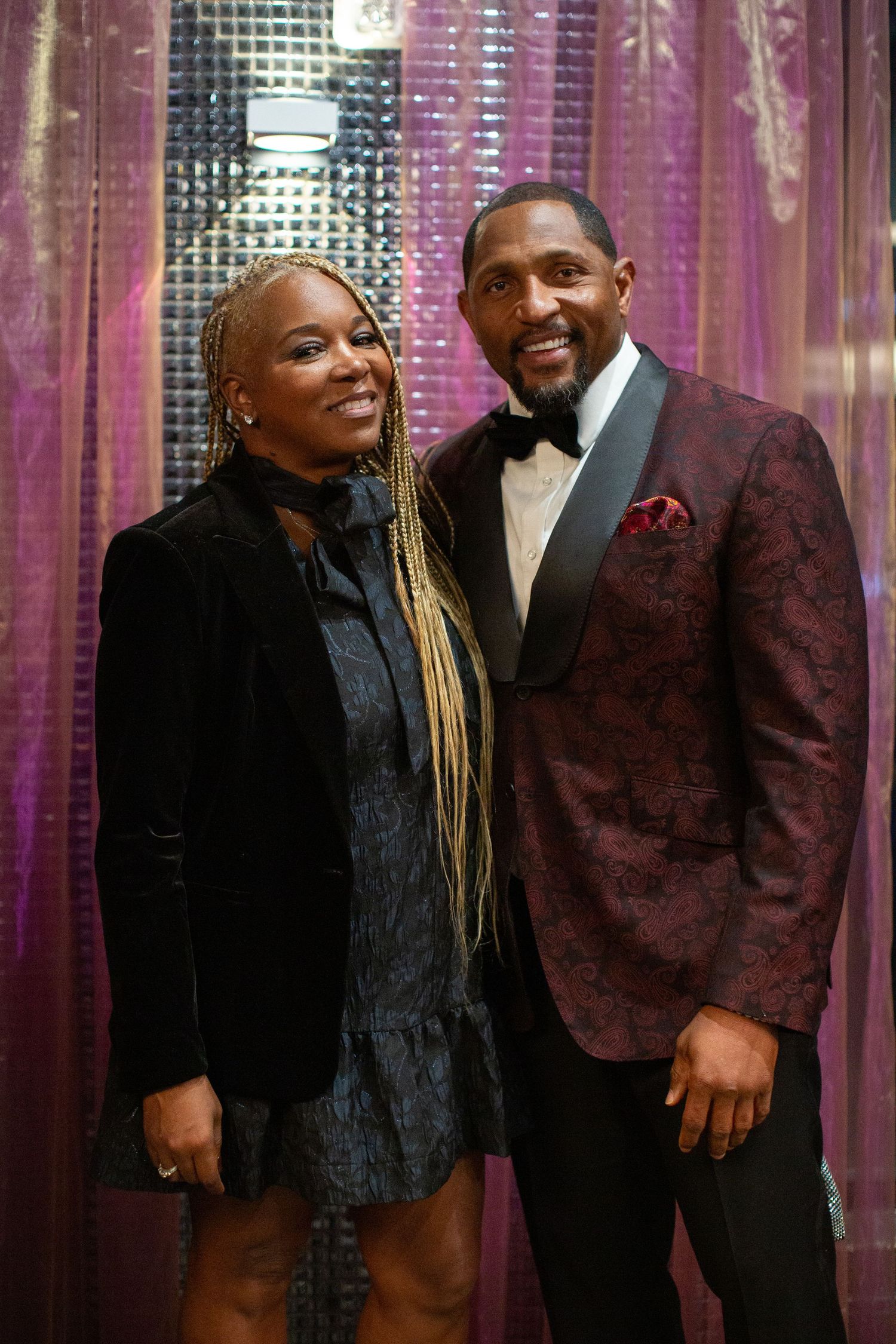 A glamorous couple poses together against a sparkly purple backdrop at an elegant evening event.