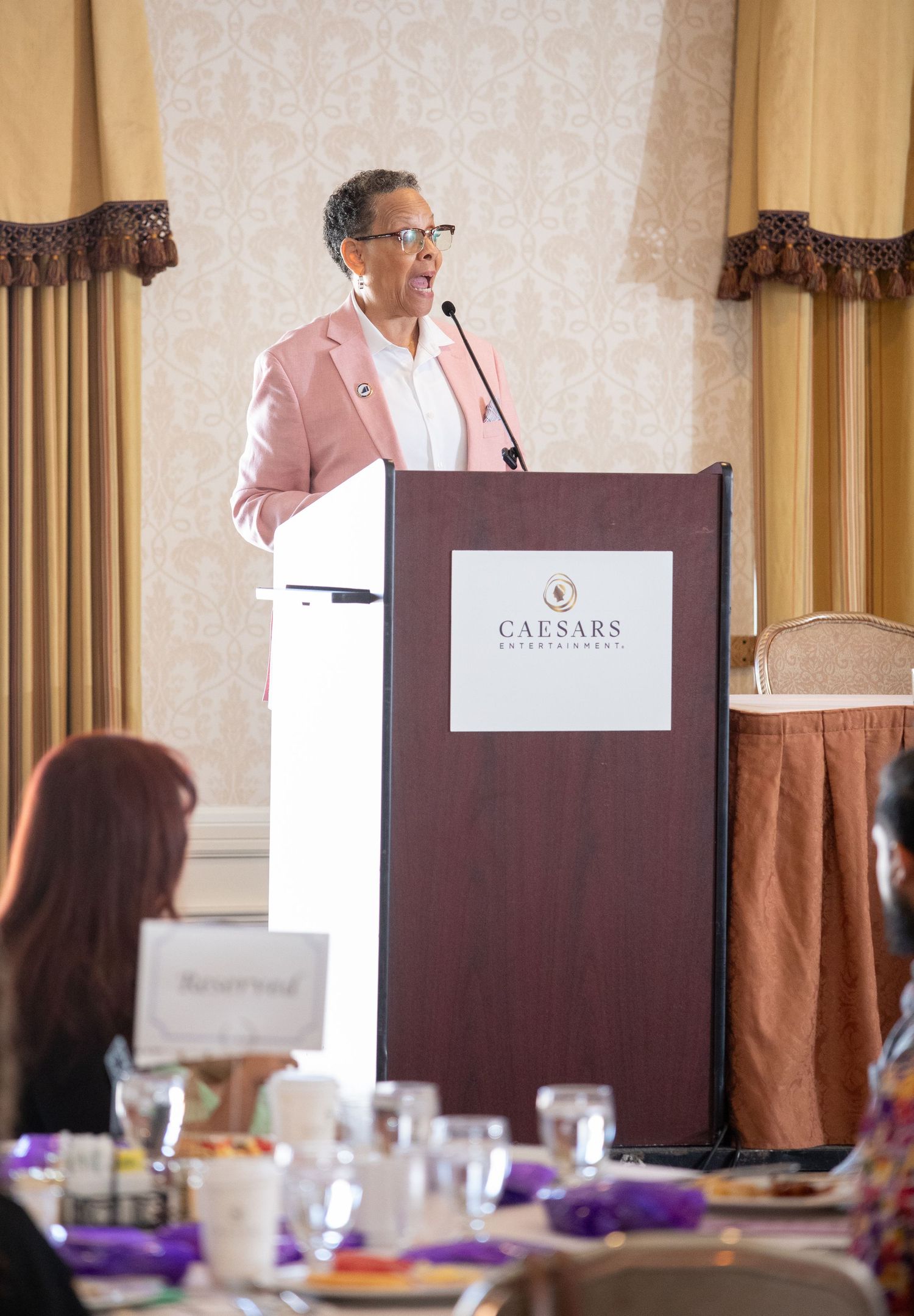 Speaker at Caesars podium wearing pink blazer addresses conference attendees in elegant banquet setting.