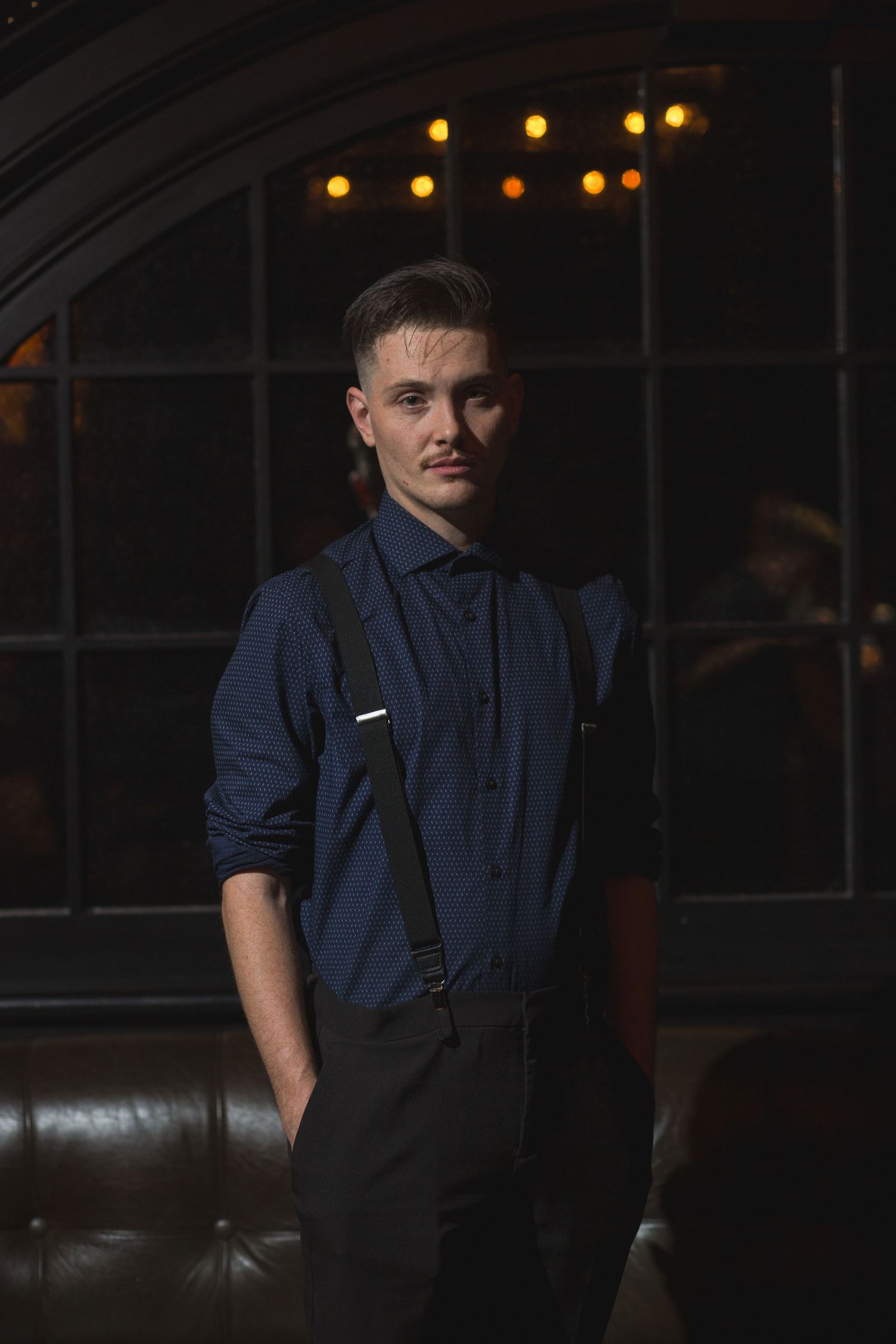A performer in suspenders and navy shirt poses in front of an arched window.