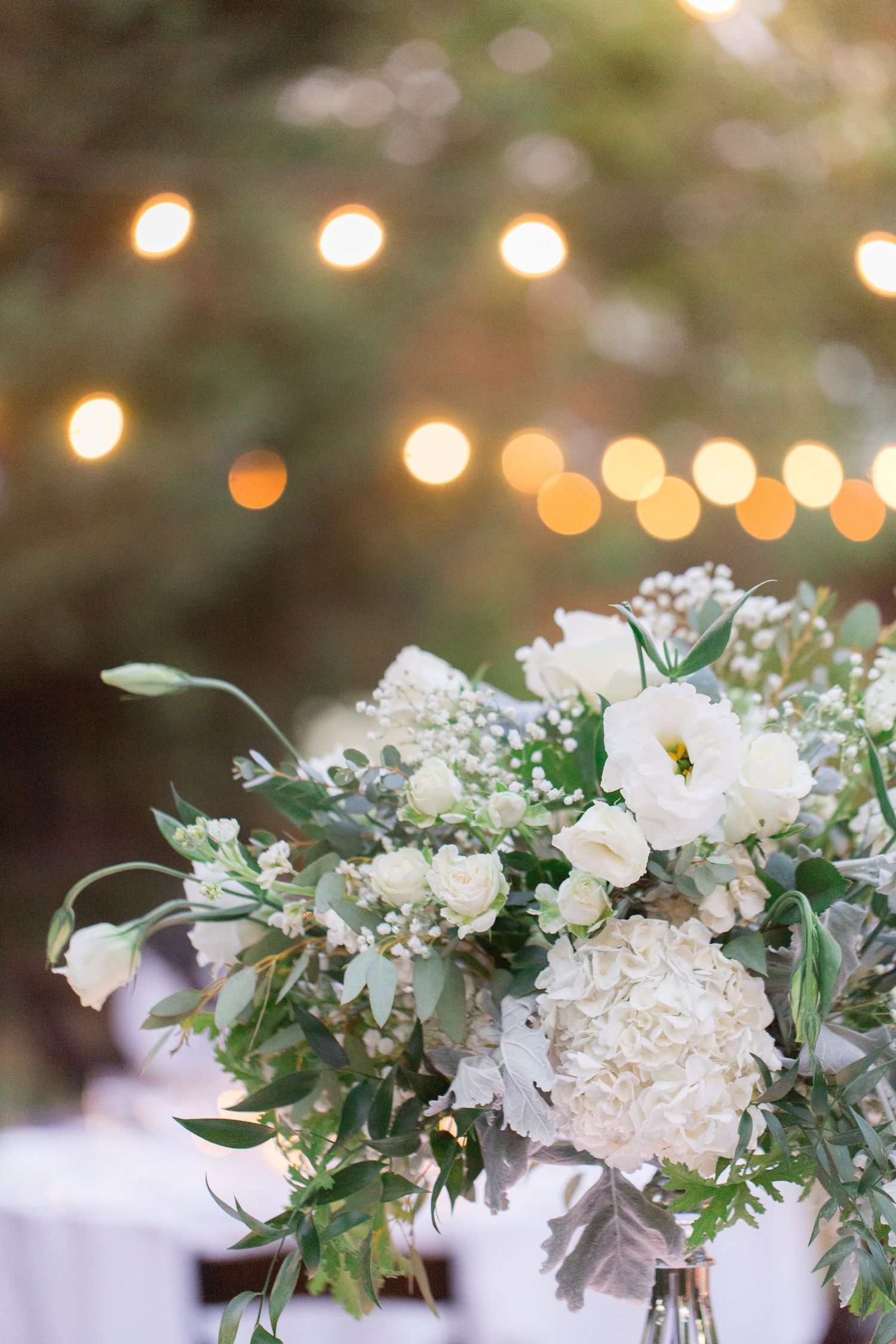 White floral centerpiece with eucalyptus against bokeh string lights background.