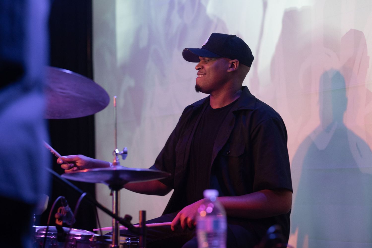 Drummer plays in low lighting with pink and purple backdrop effects.