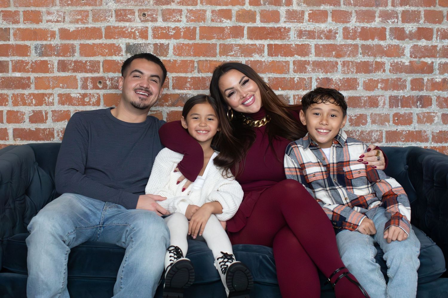 Family of four sits together on grey couch against brick wall backdrop.