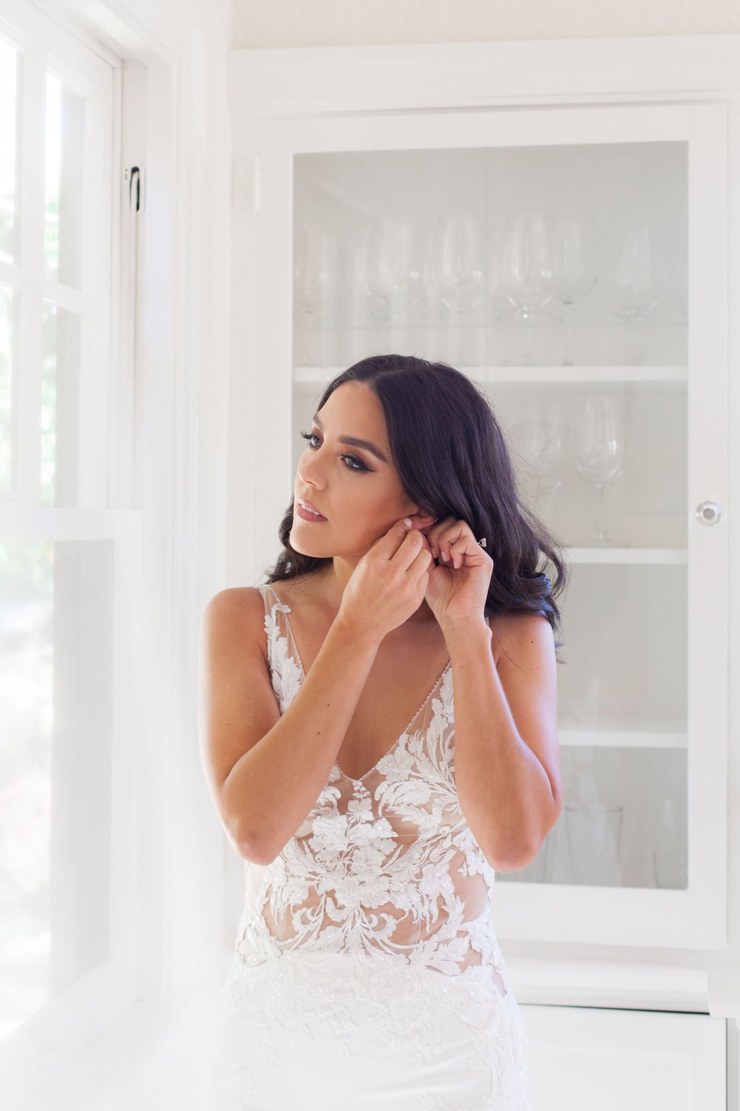 A bride in an elegant lace wedding dress adjusts her earrings in a bright white room.
