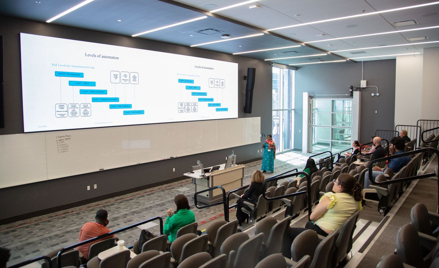 Students watch a presentation displaying timeline charts in a modern lecture theater.