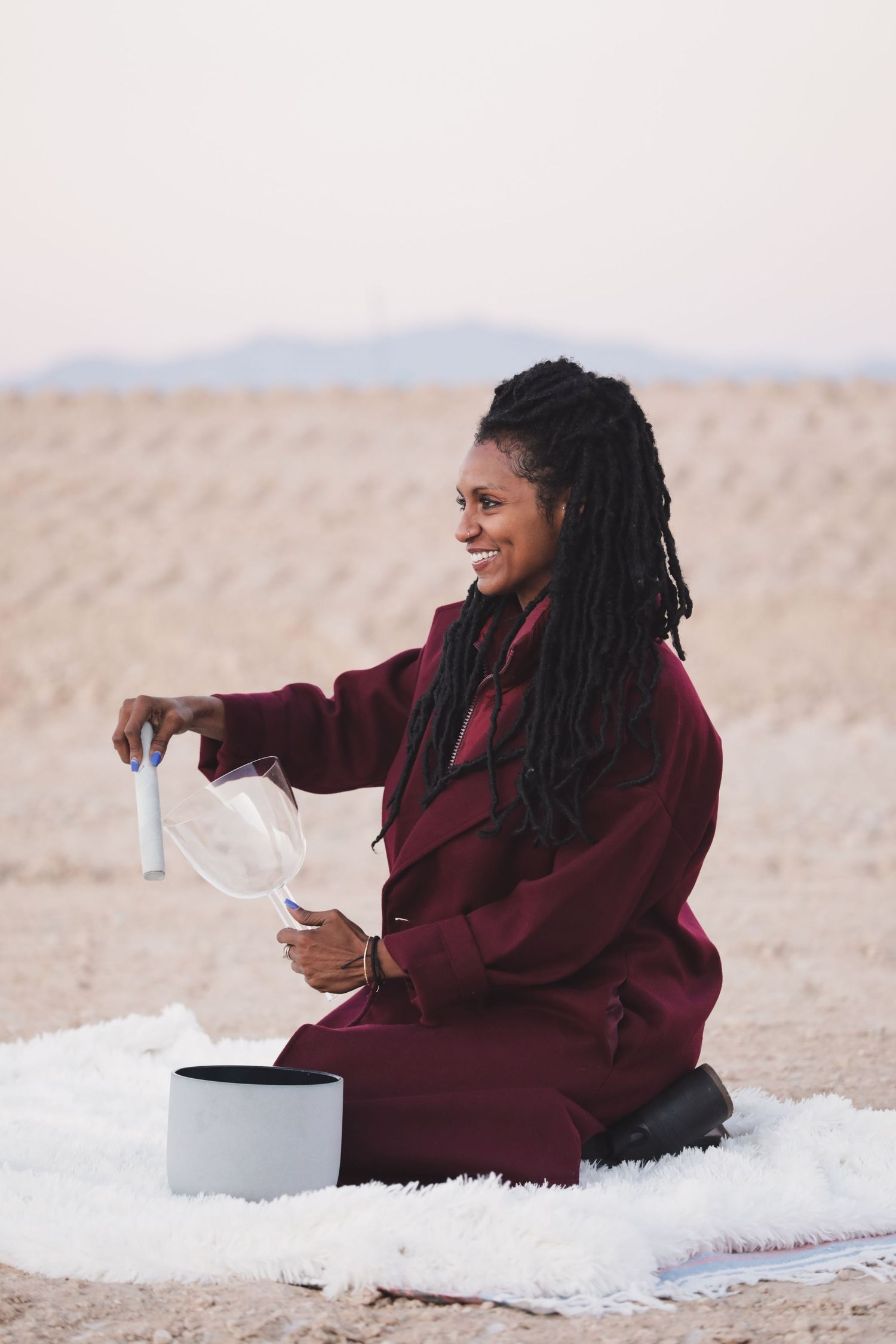 Person in burgundy clothing sitting on white blanket in desert landscape.