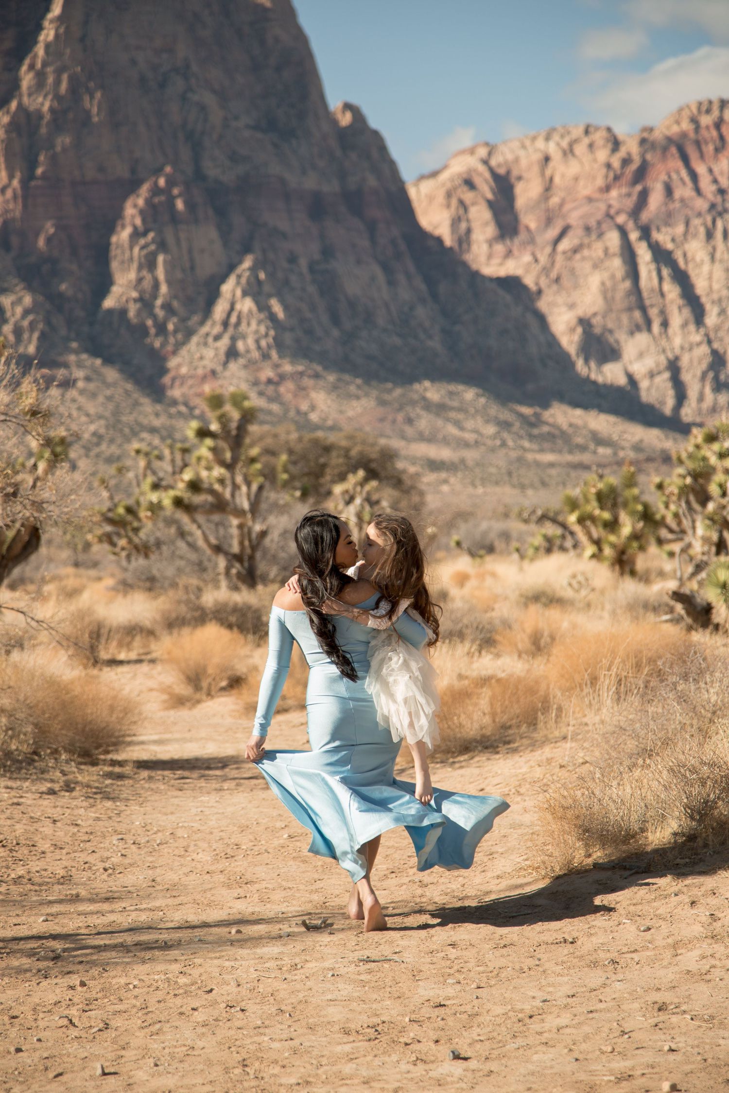 A flowing blue dress swirls on a desert trail with dramatic red rock cliffs and Joshua trees in the background.