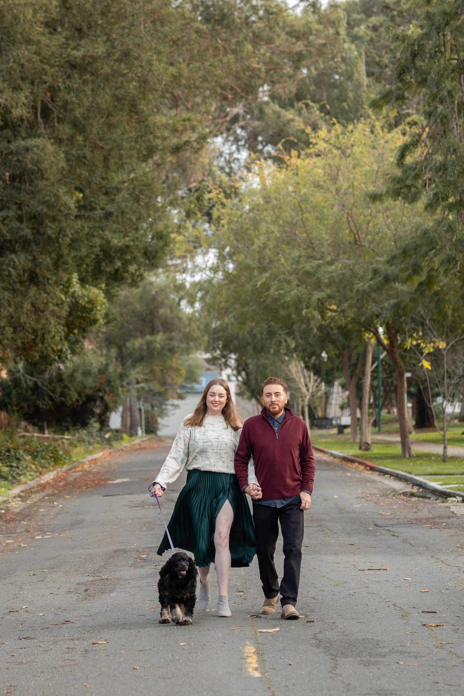 A couple walks with their black dog down a tree-lined path covered in fall leaves.