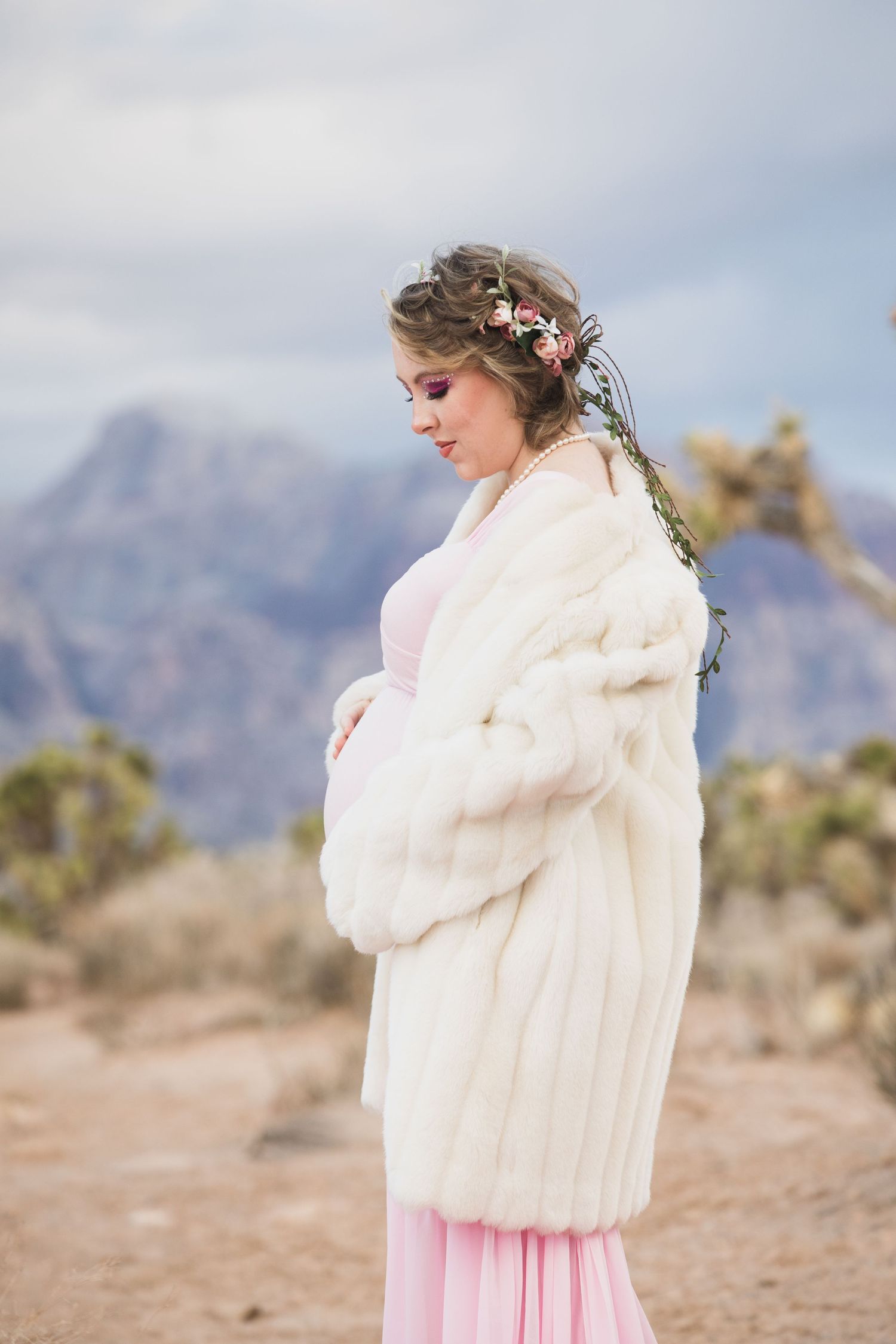 A maternity photo shoot in the desert features a pink dress and white fur coat against mountain backdrop.