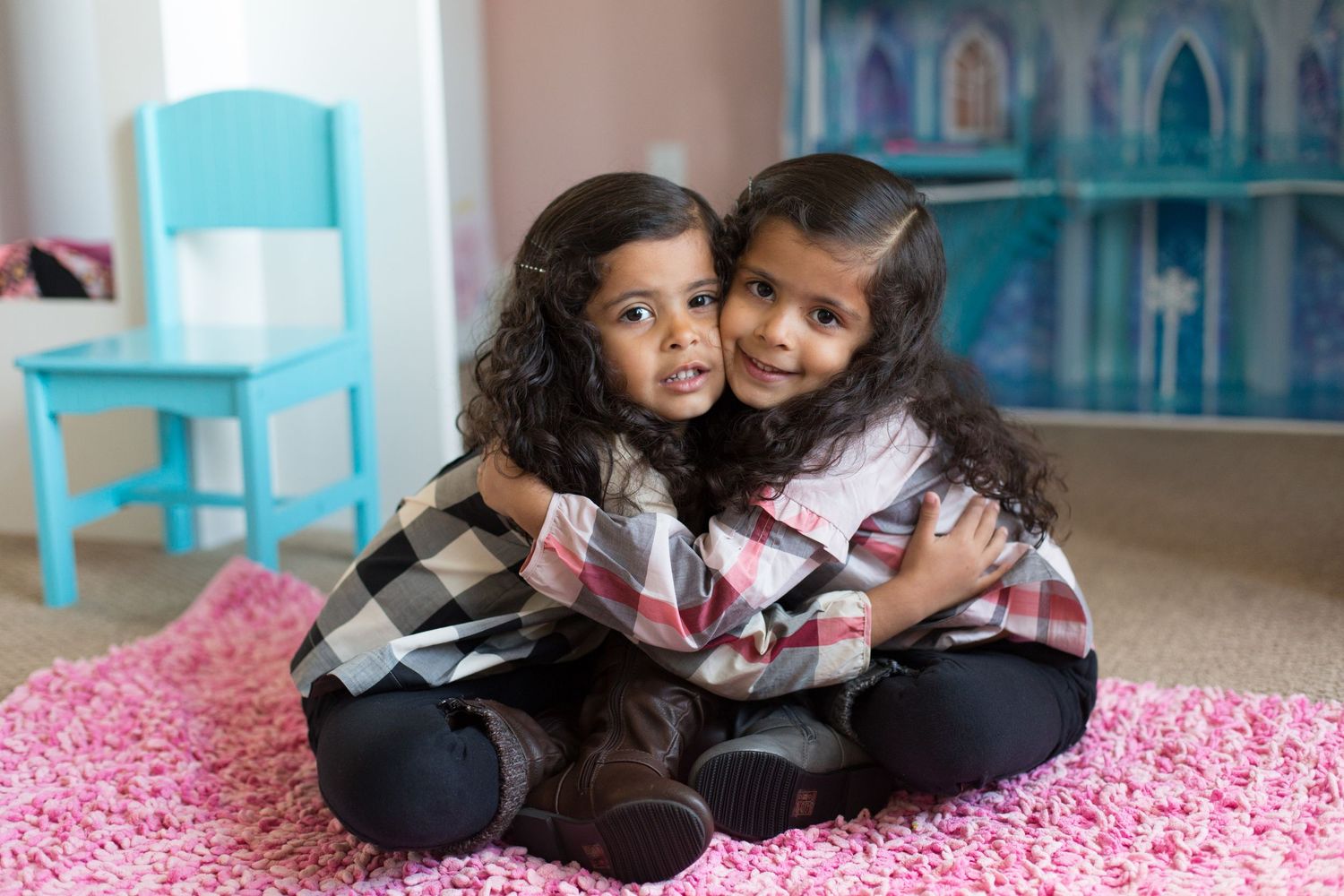 Two children embrace while sitting on pink carpet in front of blue dollhouse.