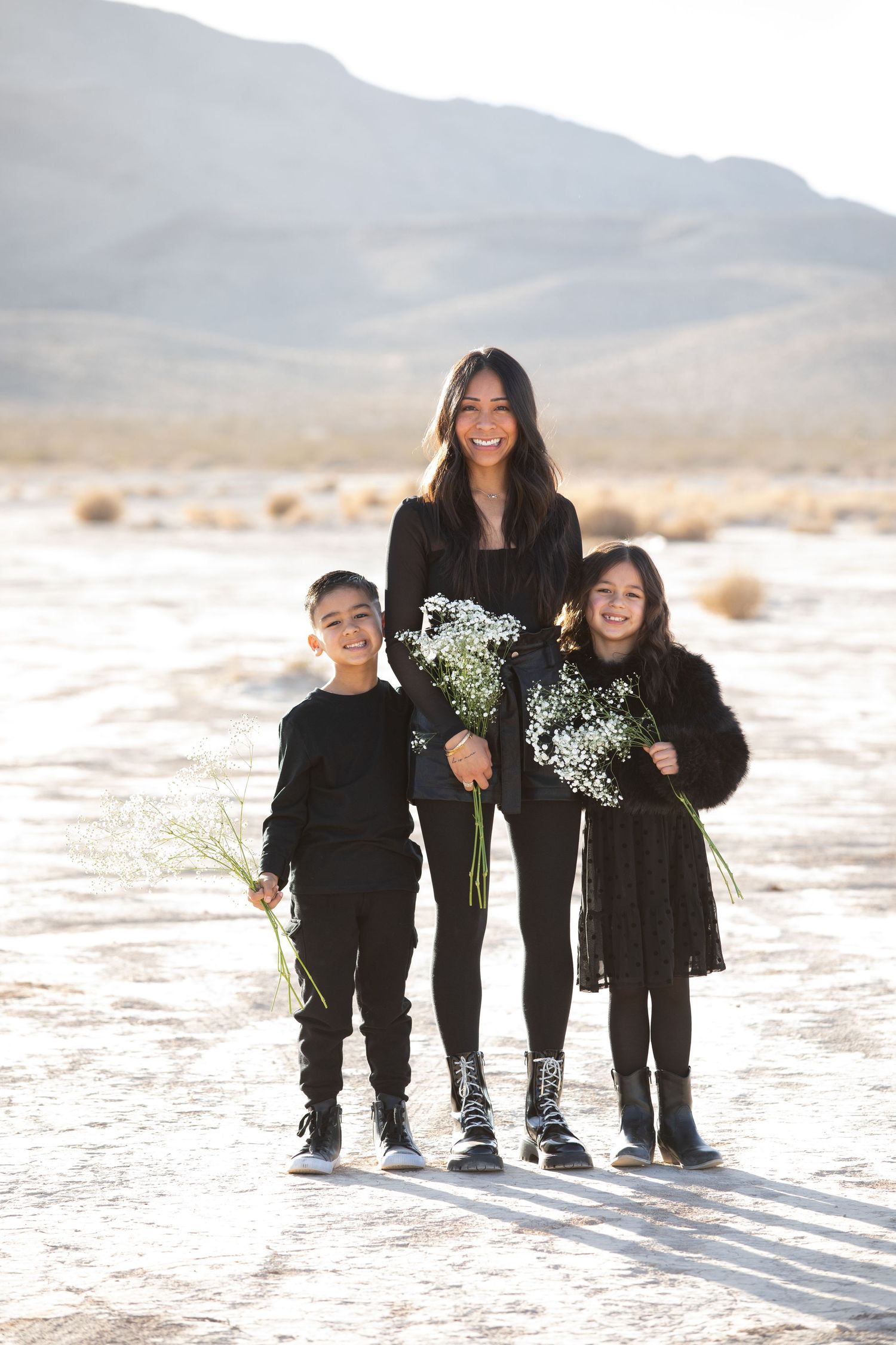 Three people in black outfits holding white flowers pose together in a desert landscape with mountains in background.