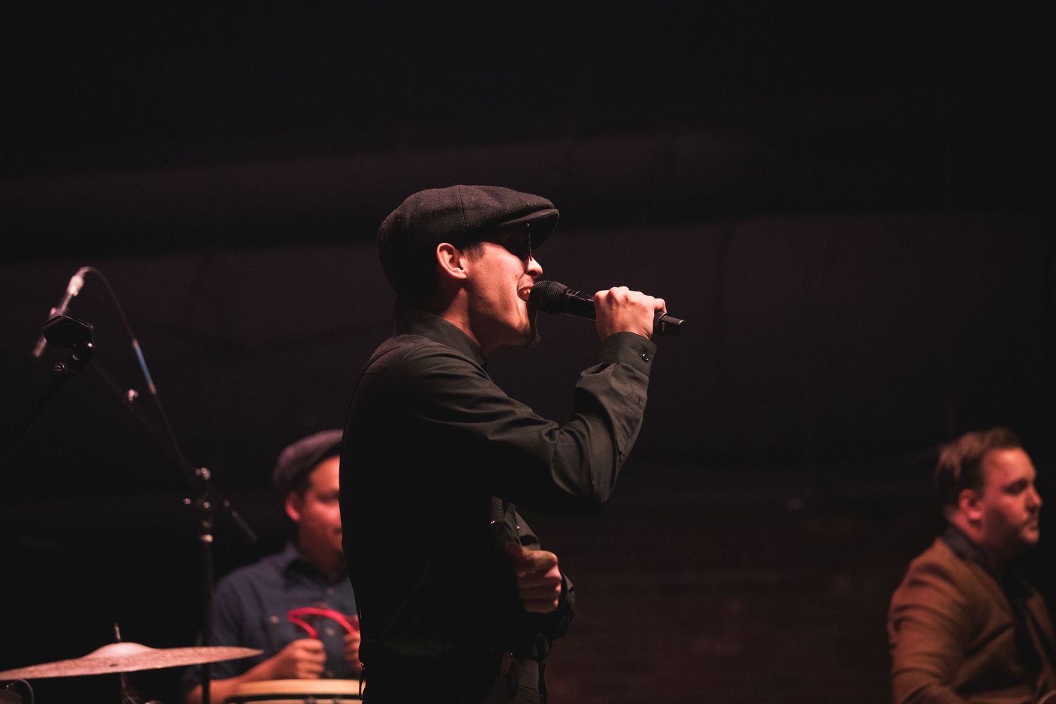 A performer in a dark hat sings into a microphone on a dimly lit stage.